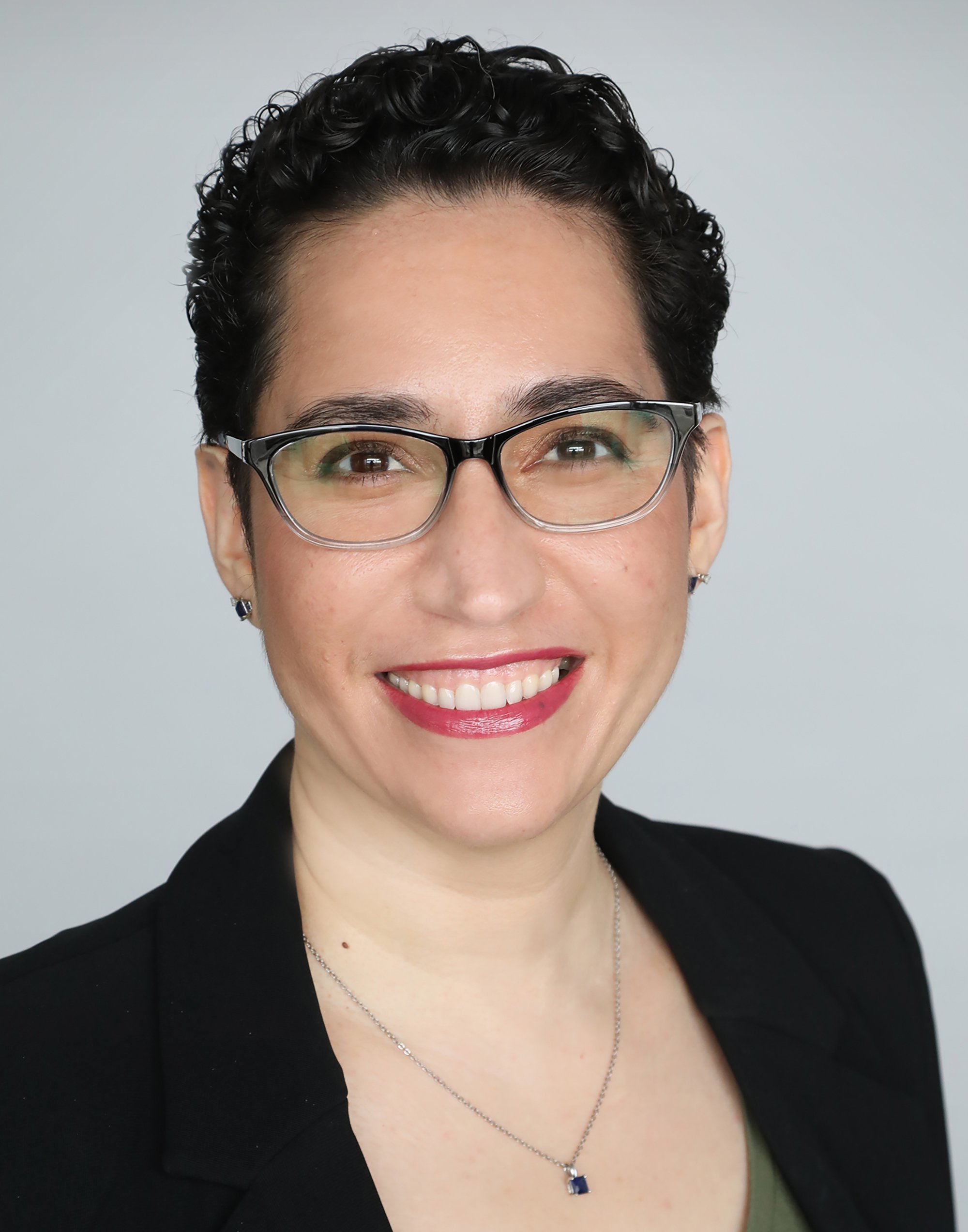 Close-up of a smiling woman with short black curly hair, wearing large eyeglasses, a black blazer, a necklace with a blue pendant, and earrings, against a plain grey background.