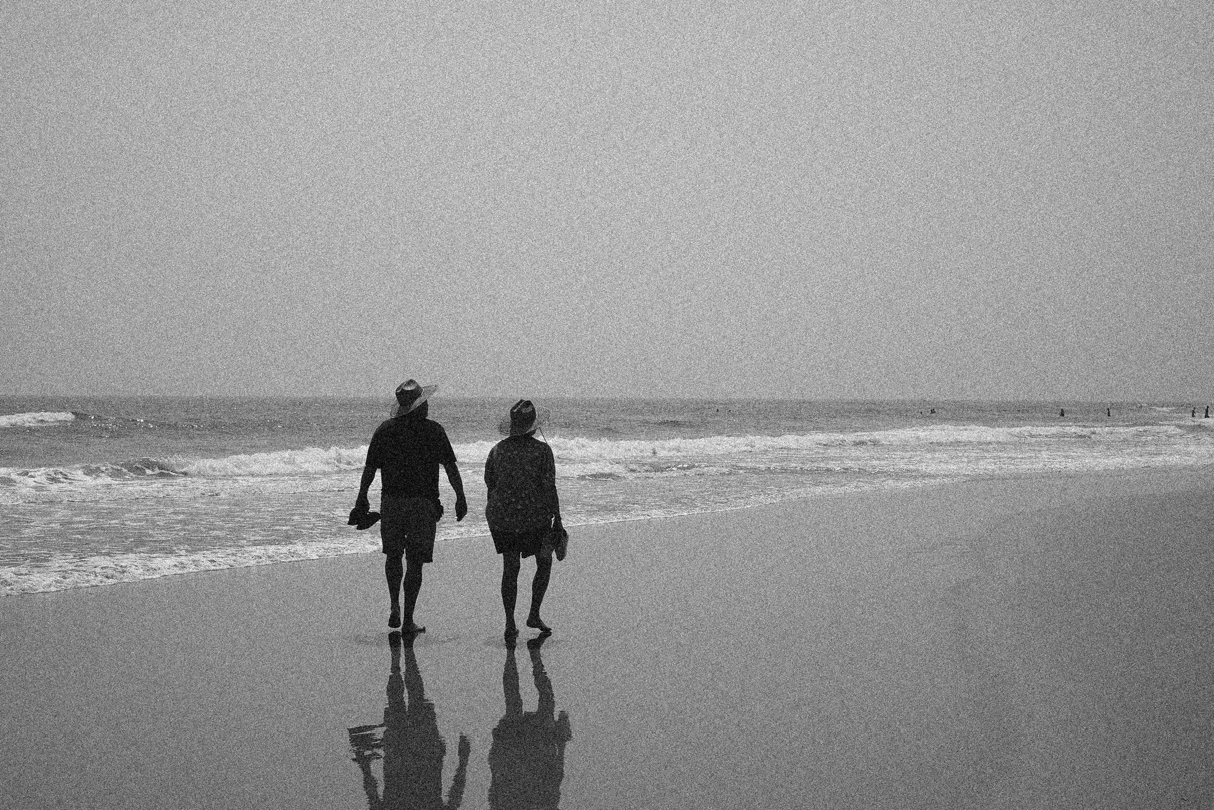 Two people holding hands walk along a beach with waves and a cloudy sky, their reflections visible on the wet sand.