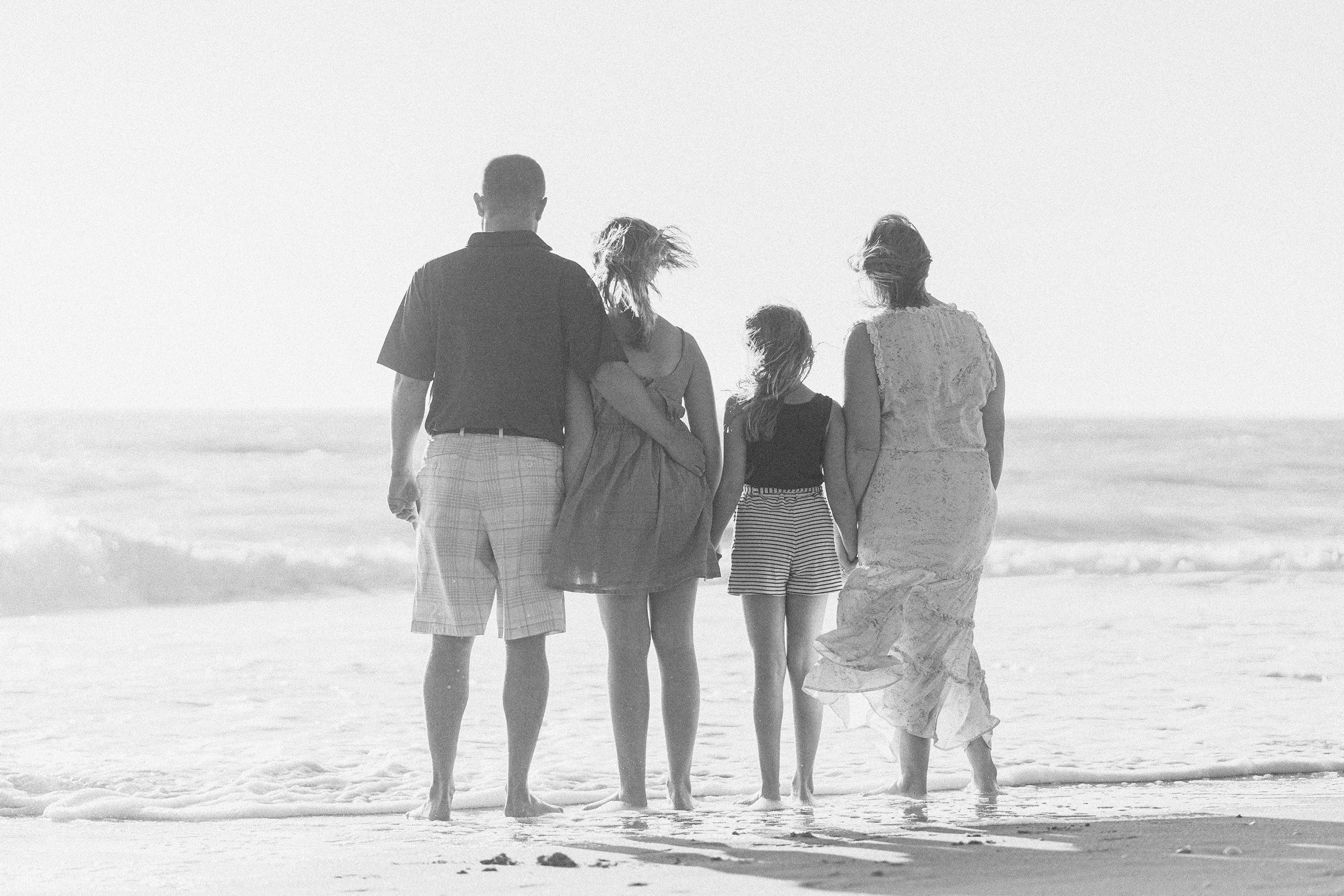 A black and white photo of five people walking on the beach, facing away from the camera, with waves in the background.