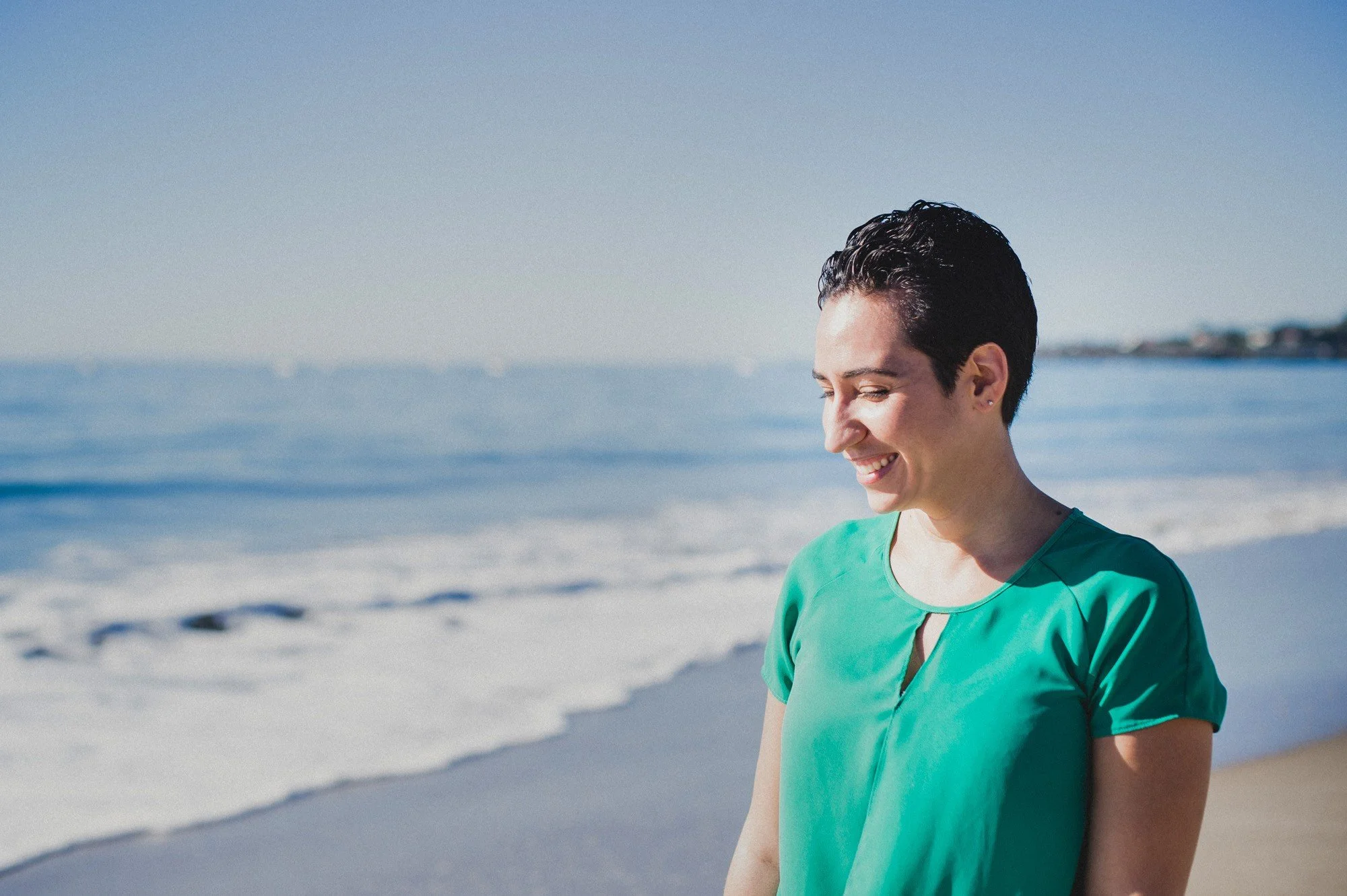 A woman with short dark hair smiling and walking on a beach with calm ocean waves and a distant shoreline under a clear blue sky.