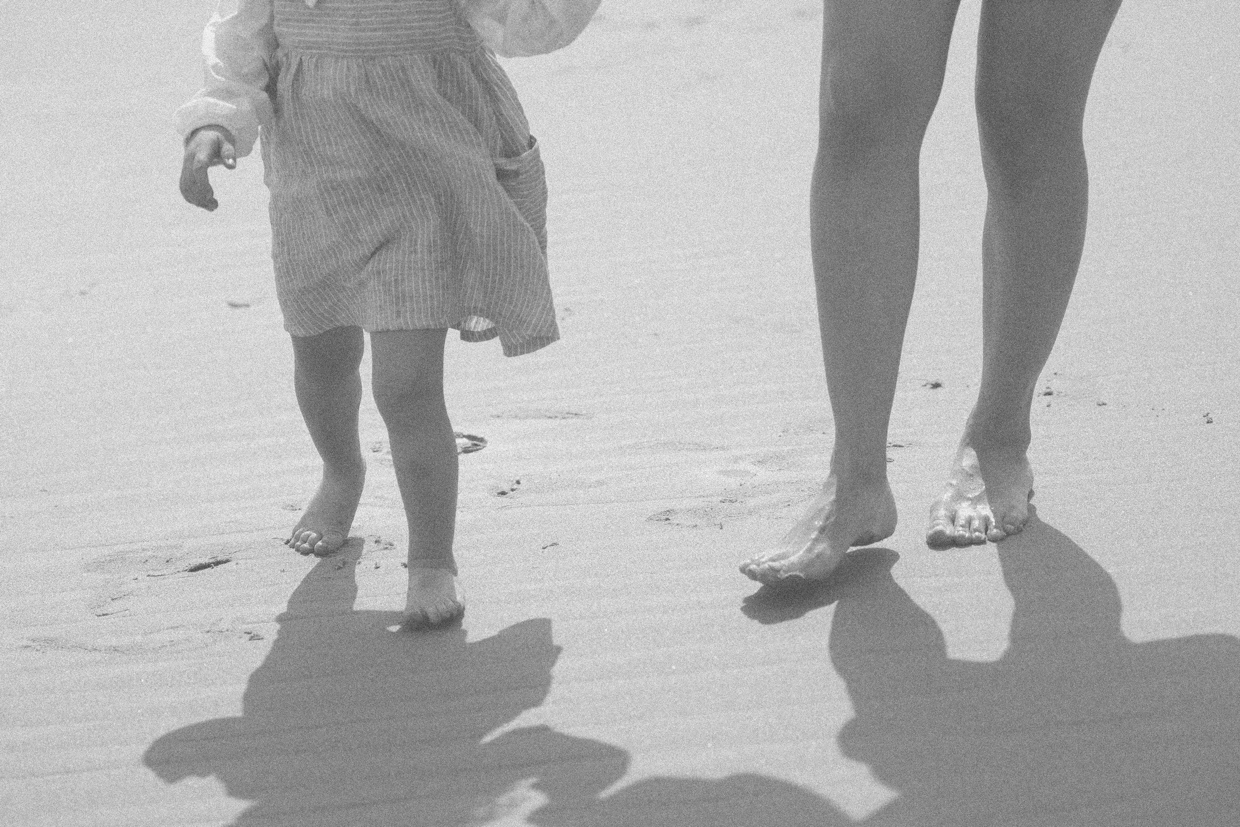 Close-up of the legs of a young child and an adult woman walking barefoot on the beach, with sand visible and shadows cast on the ground.