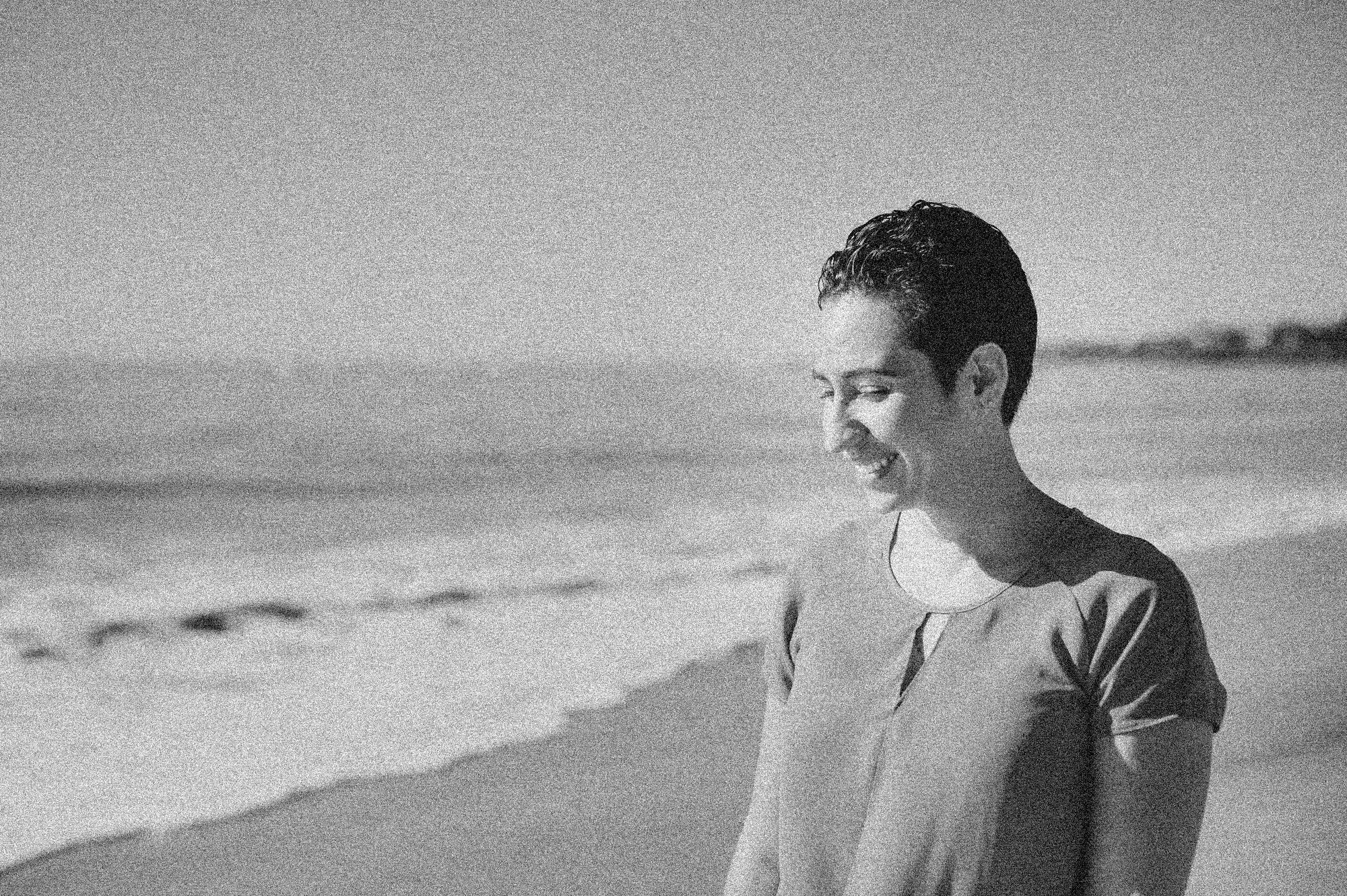 A young man smiling while walking on a beach, with the ocean and shoreline in the background.