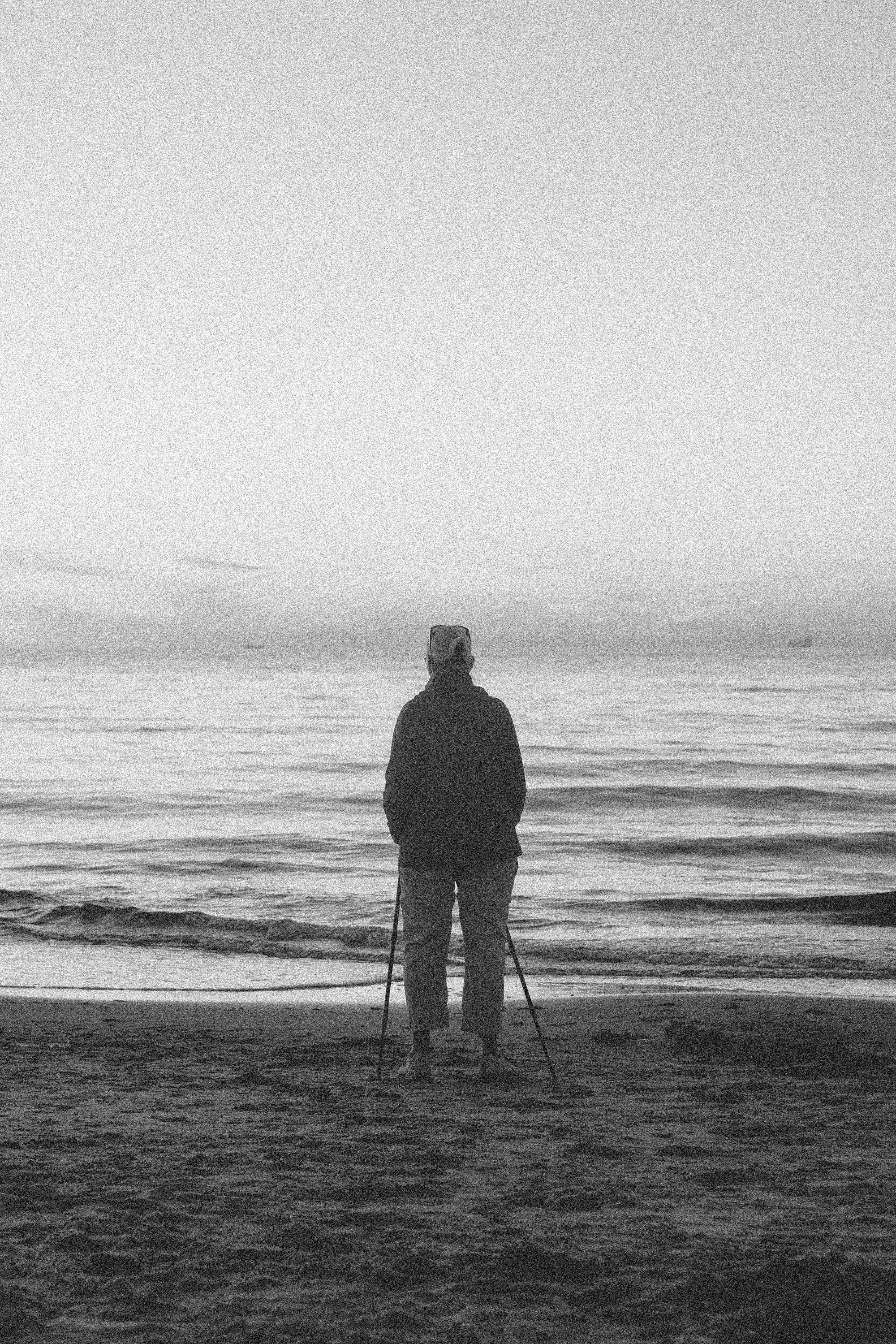 A person standing on a beach facing the ocean at dusk, holding walking sticks, with their back to the camera.