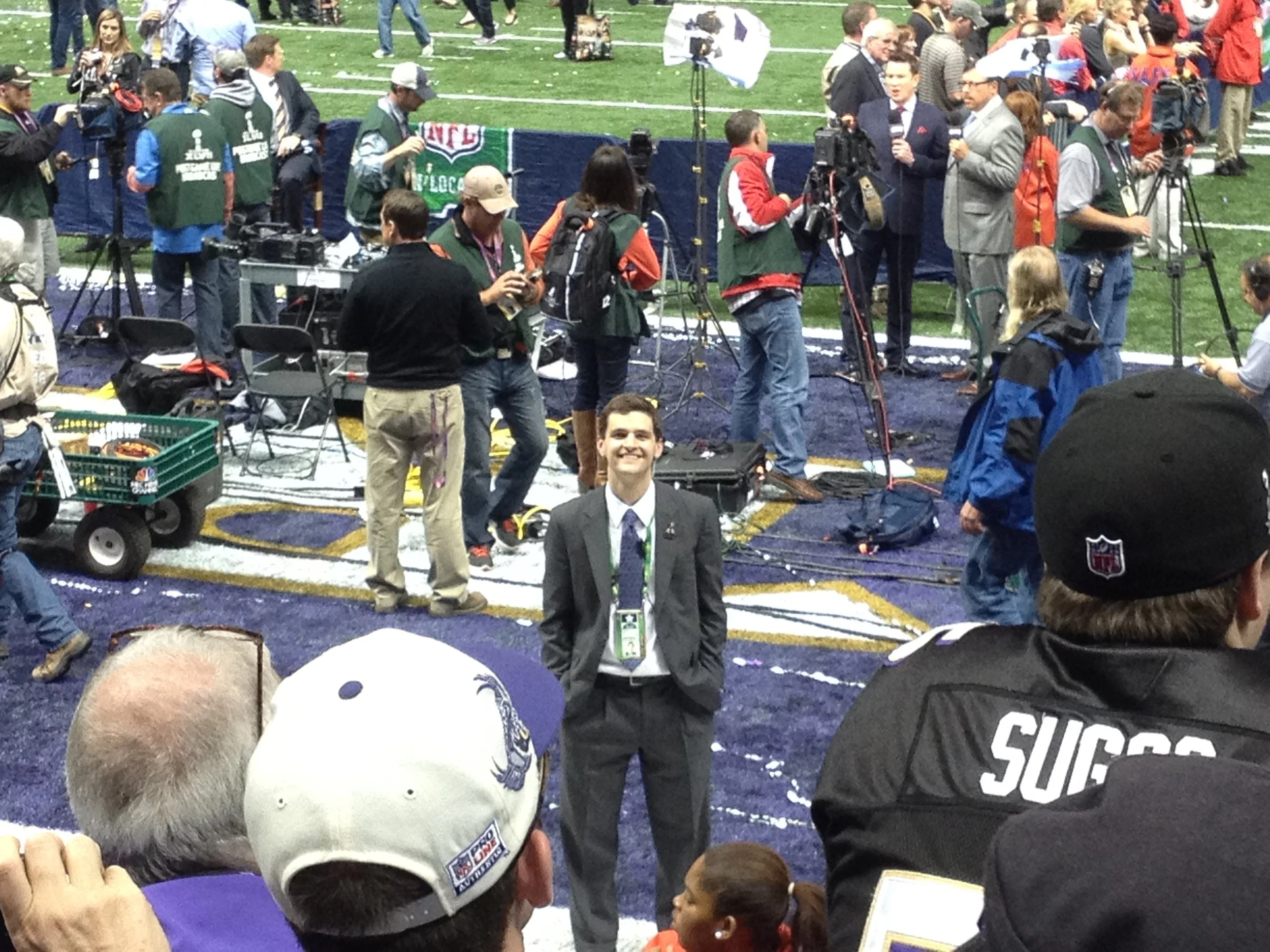 A crowded NFL press event on a football field with cameras, reporters, and security personnel, featuring a smiling Jonathan Payne in a suit and tie standing in the center.