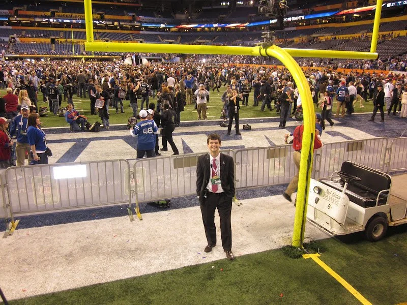 Jonathan Payne in a suit standing near a goalpost on a football field, with a large crowd of people in the background inside a stadium.