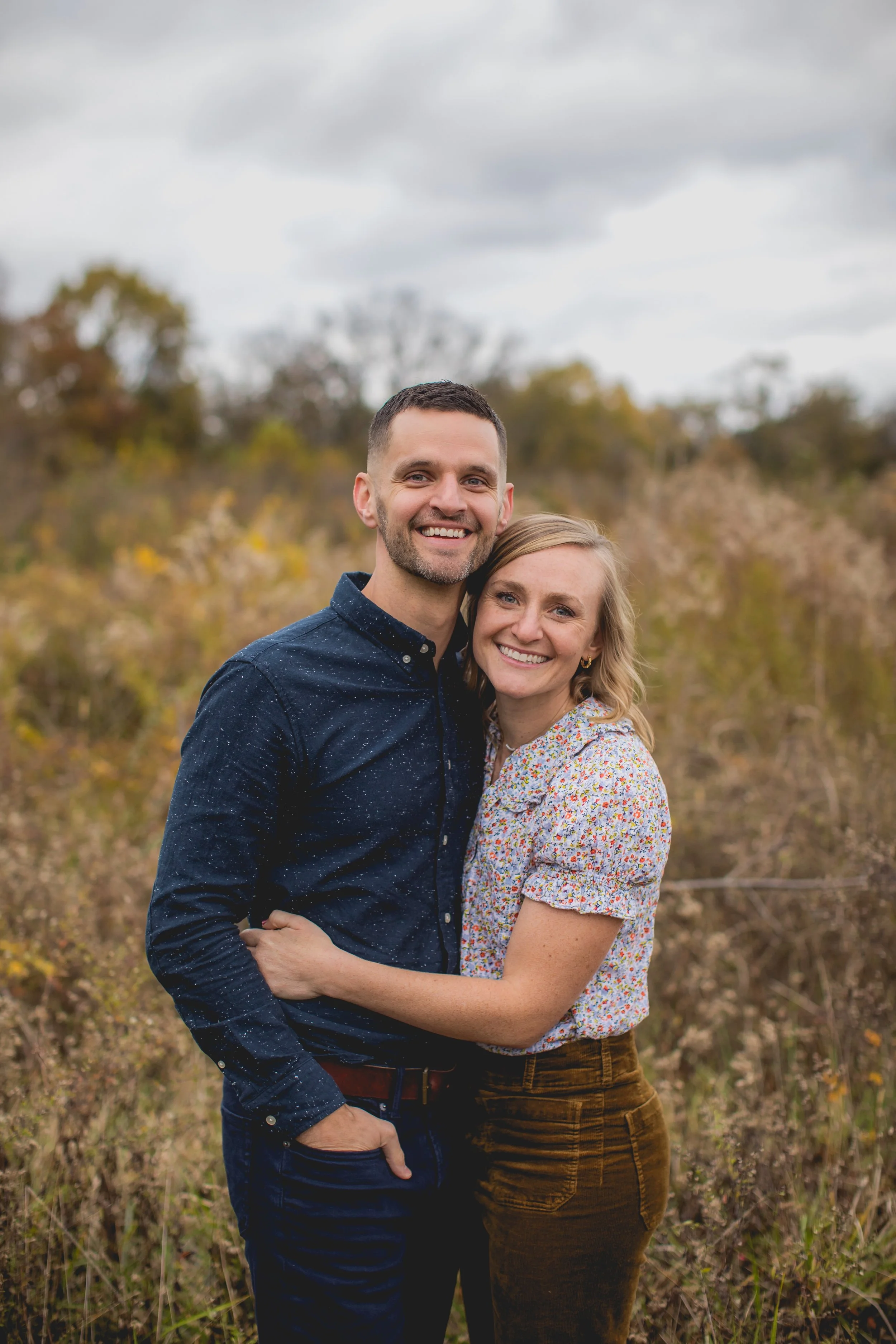 A smiling couple standing close together outdoors in a field with autumn foliage, under a cloudy sky.