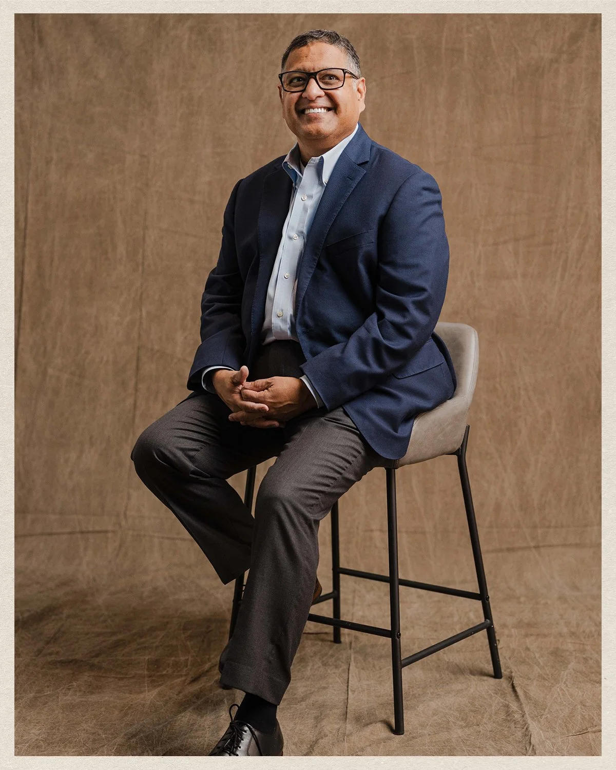 Neal Patel in a navy blazer and gray pants sitting on a barstool against a brown background, smiling at the camera.