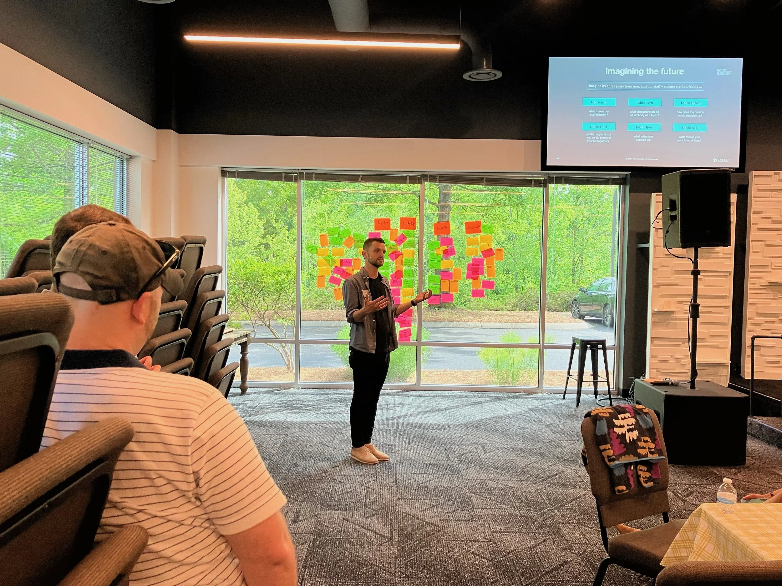 A man is giving a presentation in a room with large windows and a backdrop covered in colorful sticky notes. Several audience members are seated, some taking notes. A large screen displays a slide titled 'Imagining the Future' with various bullet points.