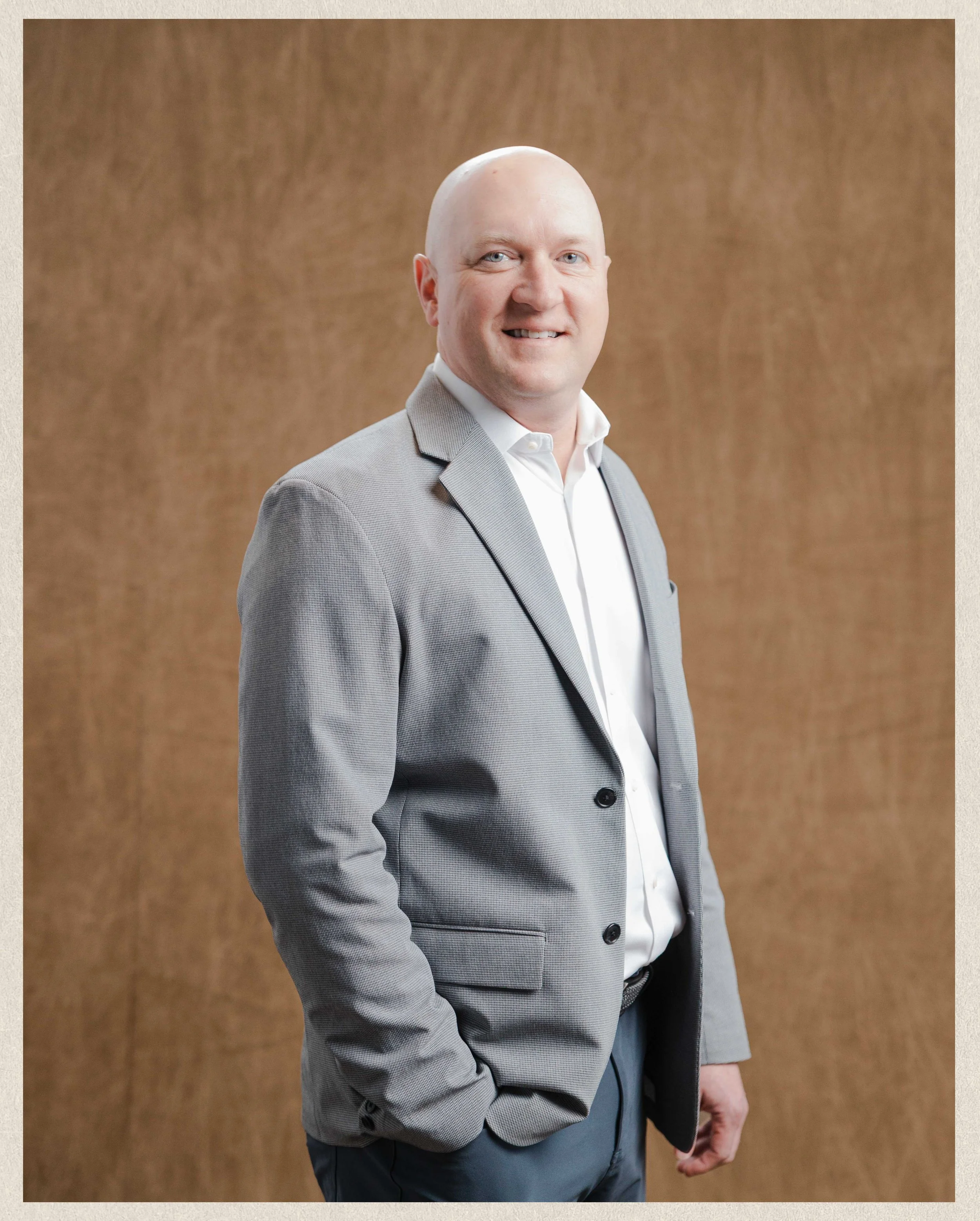 Portrait of a bald man in a light gray suit and white shirt, smiling, standing against a wooden background.