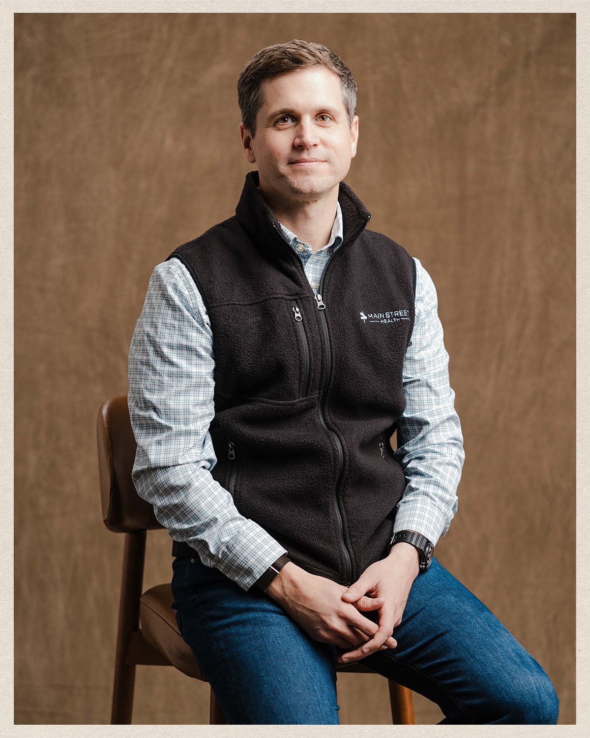A man sitting on a wooden chair against a plain brown background. He has short brown hair and is wearing a light checkered button-up shirt, a black zip-up vest with a logo that reads 'Main Street', and dark blue jeans. His hands are clasped together on his lap, and he has a neutral expression.