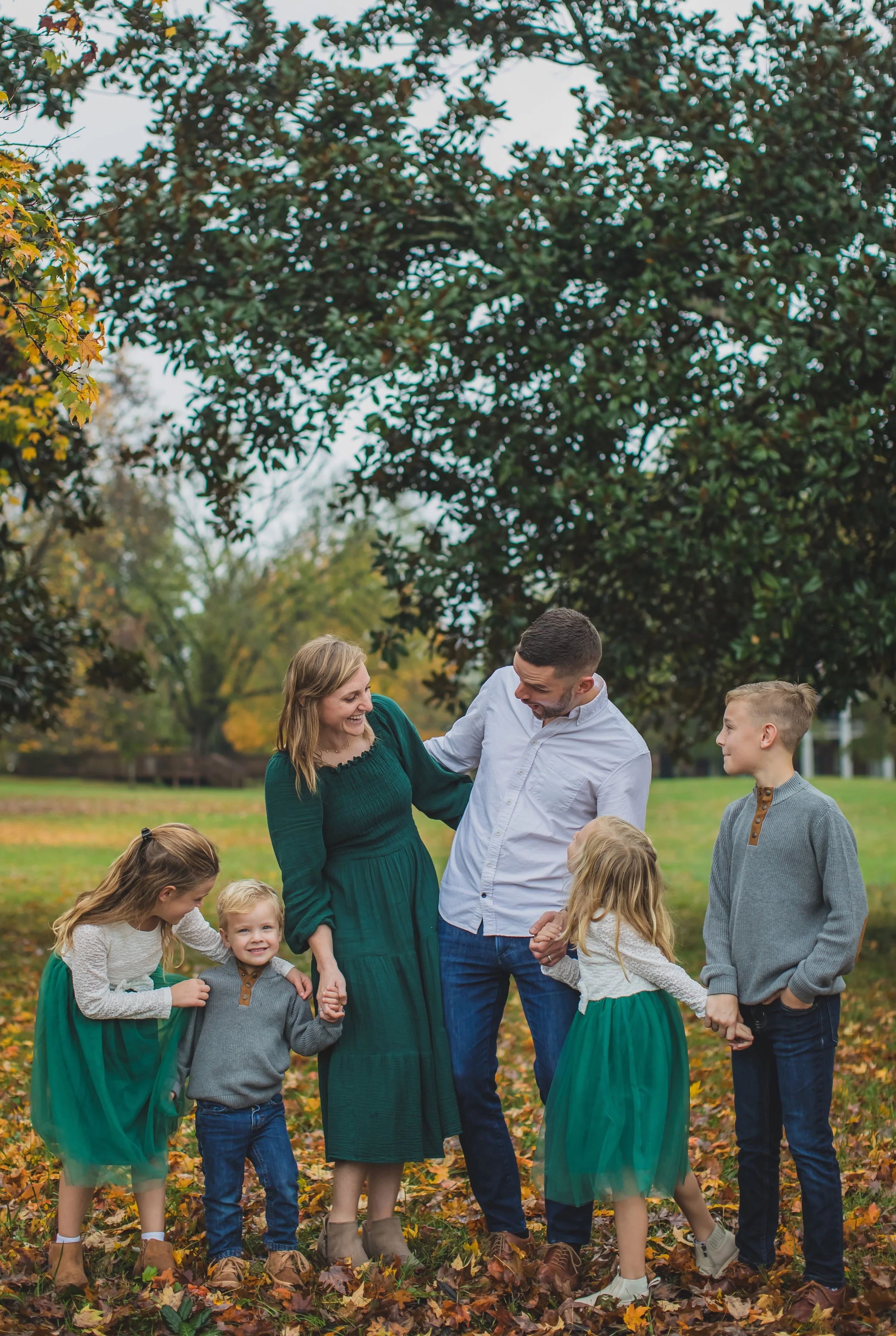 A family of six, including two adults and four children, standing outdoors in a park with fallen leaves, surrounded by trees with green and autumn-colored foliage. The family is smiling and holding hands, dressed in fall clothing.