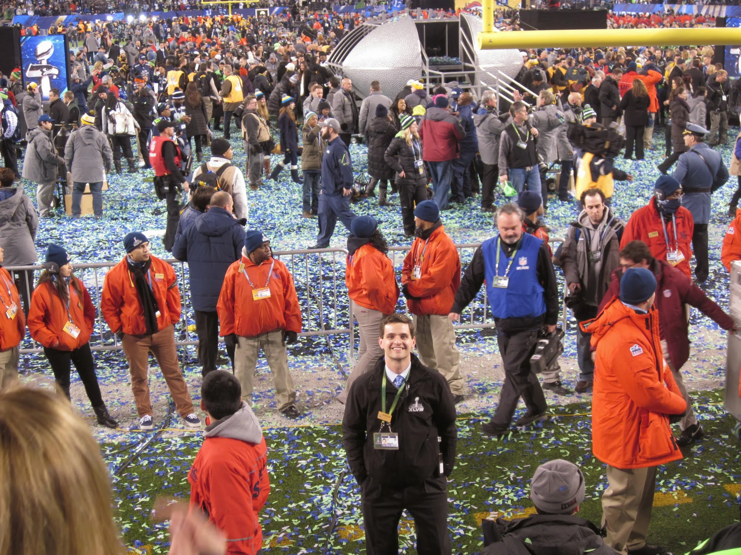 Scene from a sports event with many people, some wearing orange jackets and others with lanyards. Confetti on the ground indicates celebration, possibly after a game or medal ceremony.