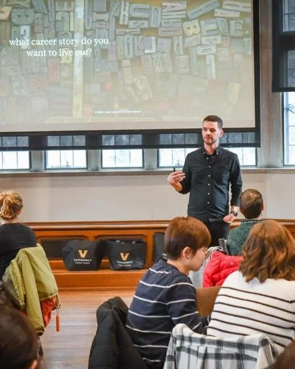 A man giving a presentation to a group of people seated at tables in a classroom or conference room. There is a large screen behind him displaying a slide with labeled building blocks and the text, 'what career story do you want to live out?' The audience appears engaged, and the room has large windows letting in natural light.
