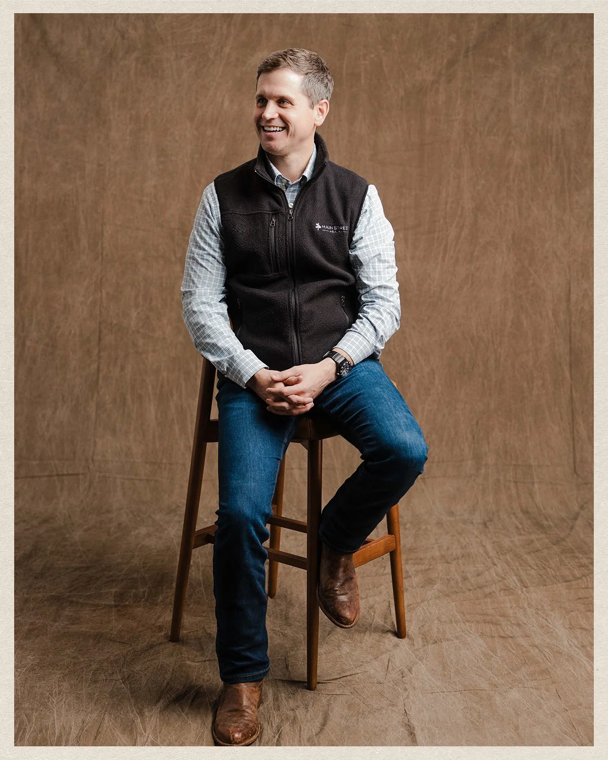 A man sitting on a wooden stool in front of a plain brown backdrop, wearing a black vest, long-sleeve plaid shirt, jeans, and cowboy boots, smiling and looking to the left.