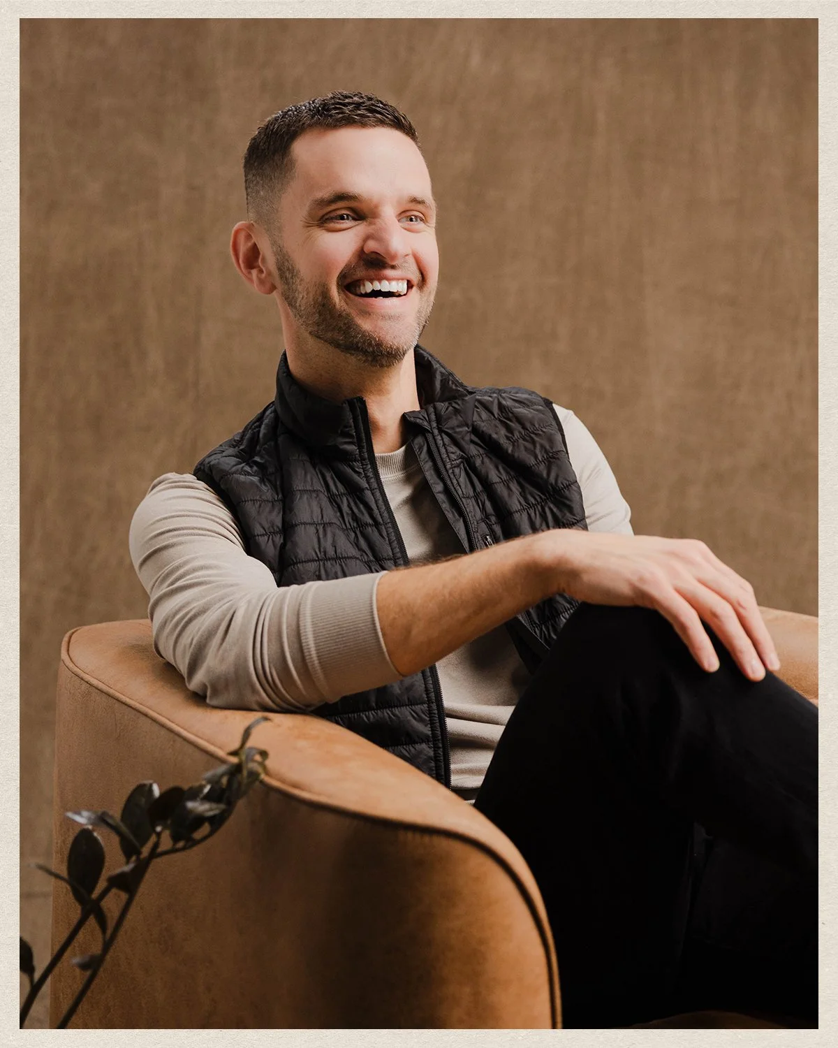 Jonathan Payne smiling man sitting on a tan armchair, wearing a black puffer vest over a beige long-sleeve shirt, with a neutral background.