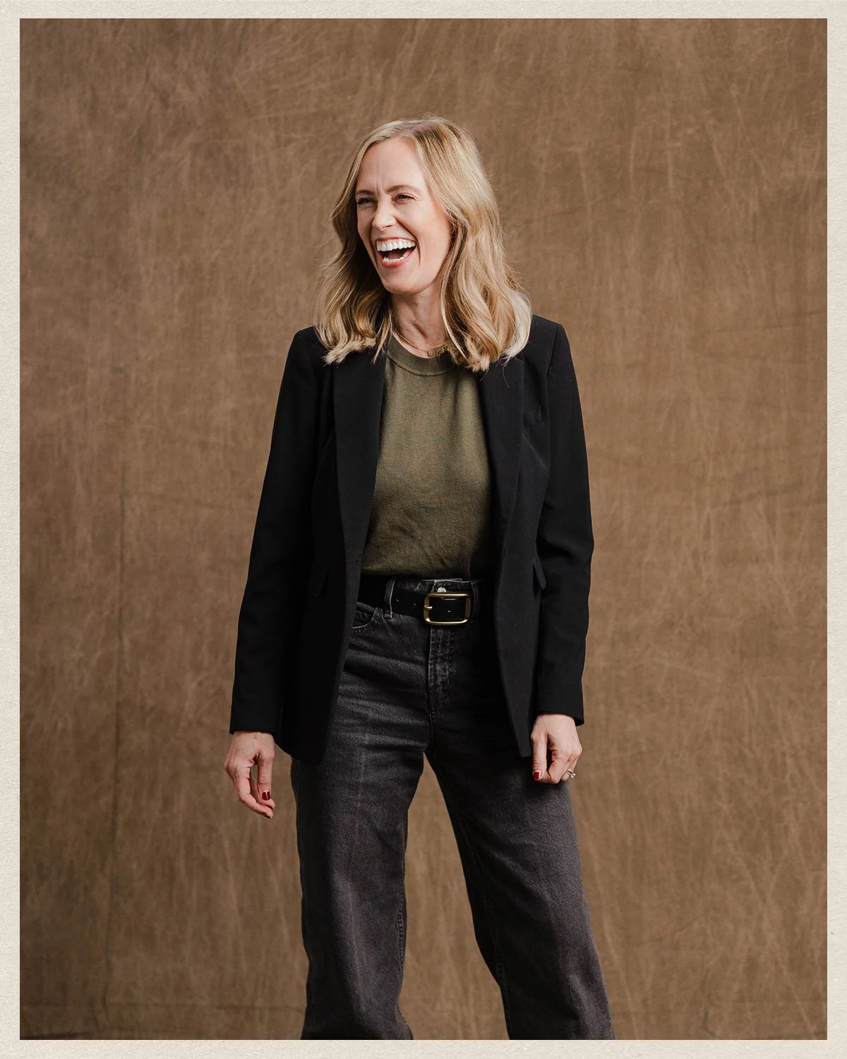 Alicia Wilson with blonde hair laughing or smiling, wearing a black blazer over an olive-green shirt and dark jeans, standing against a brown textured background.