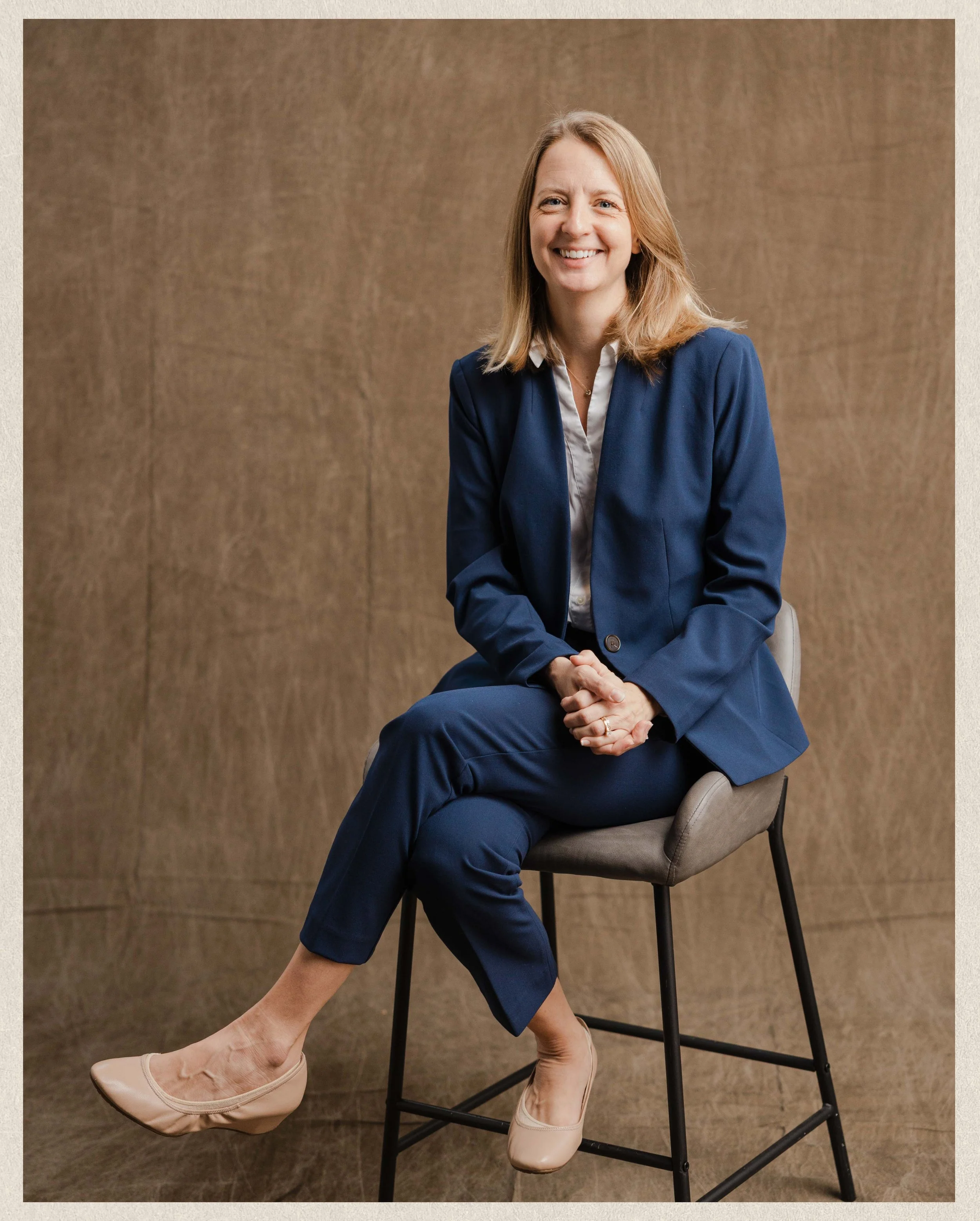 A woman in a navy blue suit with beige heels sitting on a stool, smiling, with a brown textured background.