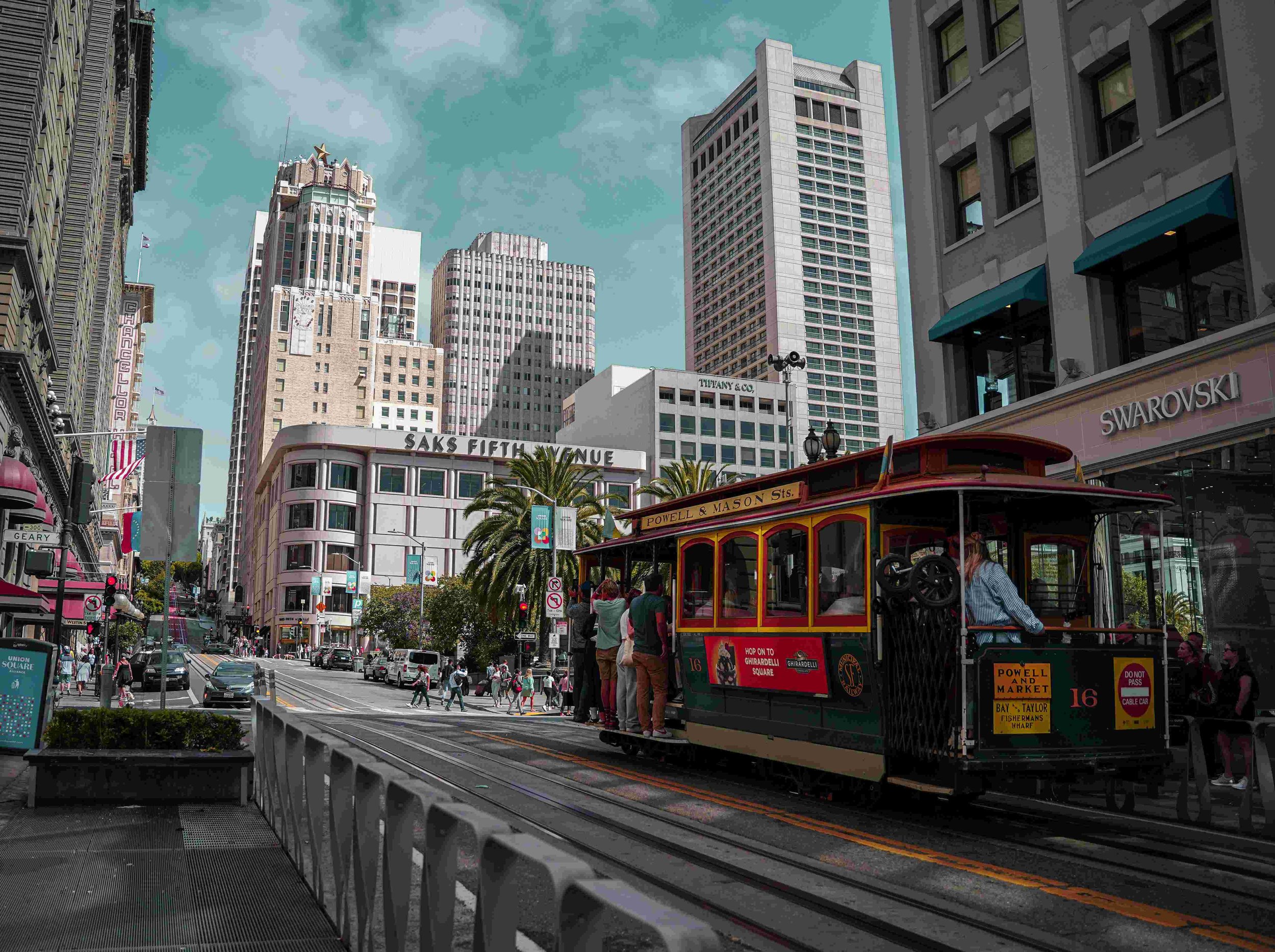 A street scene in a city with tall buildings, a trolley car on tracks, cars, and pedestrians, including a man in a blue shirt riding the trolley, under a partly cloudy sky.