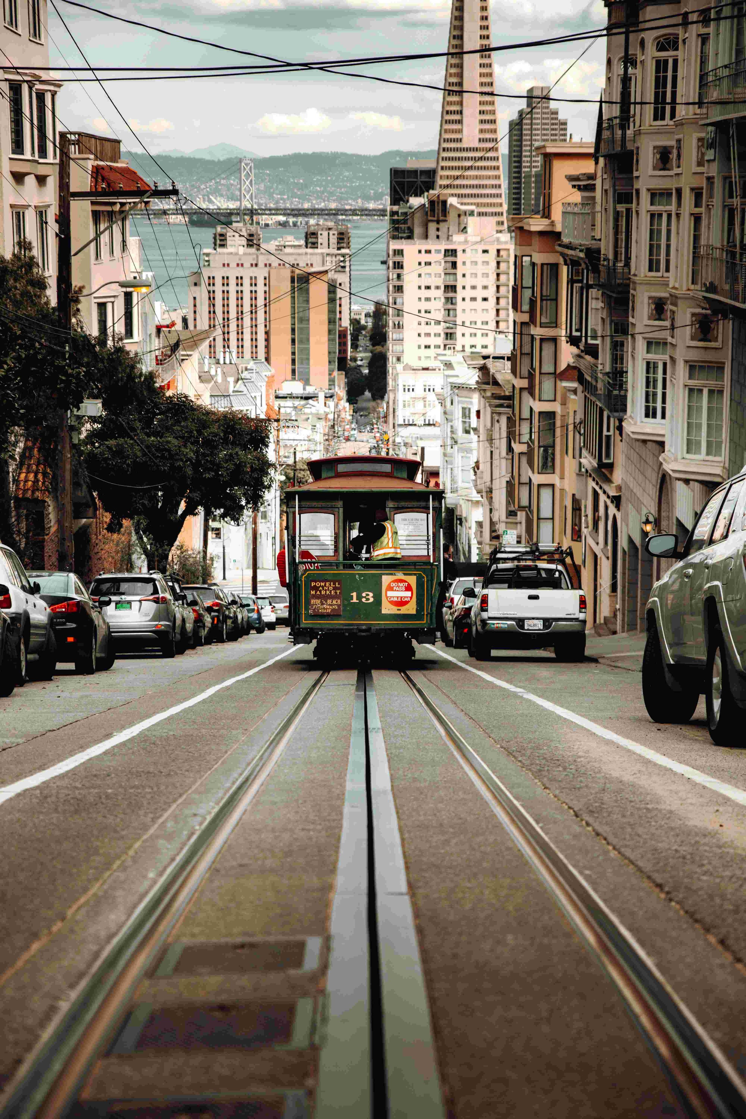 A cable car traveling down a hilly street in San Francisco with parked cars on both sides and buildings on either side, leading toward a body of water and a bridge in the background.