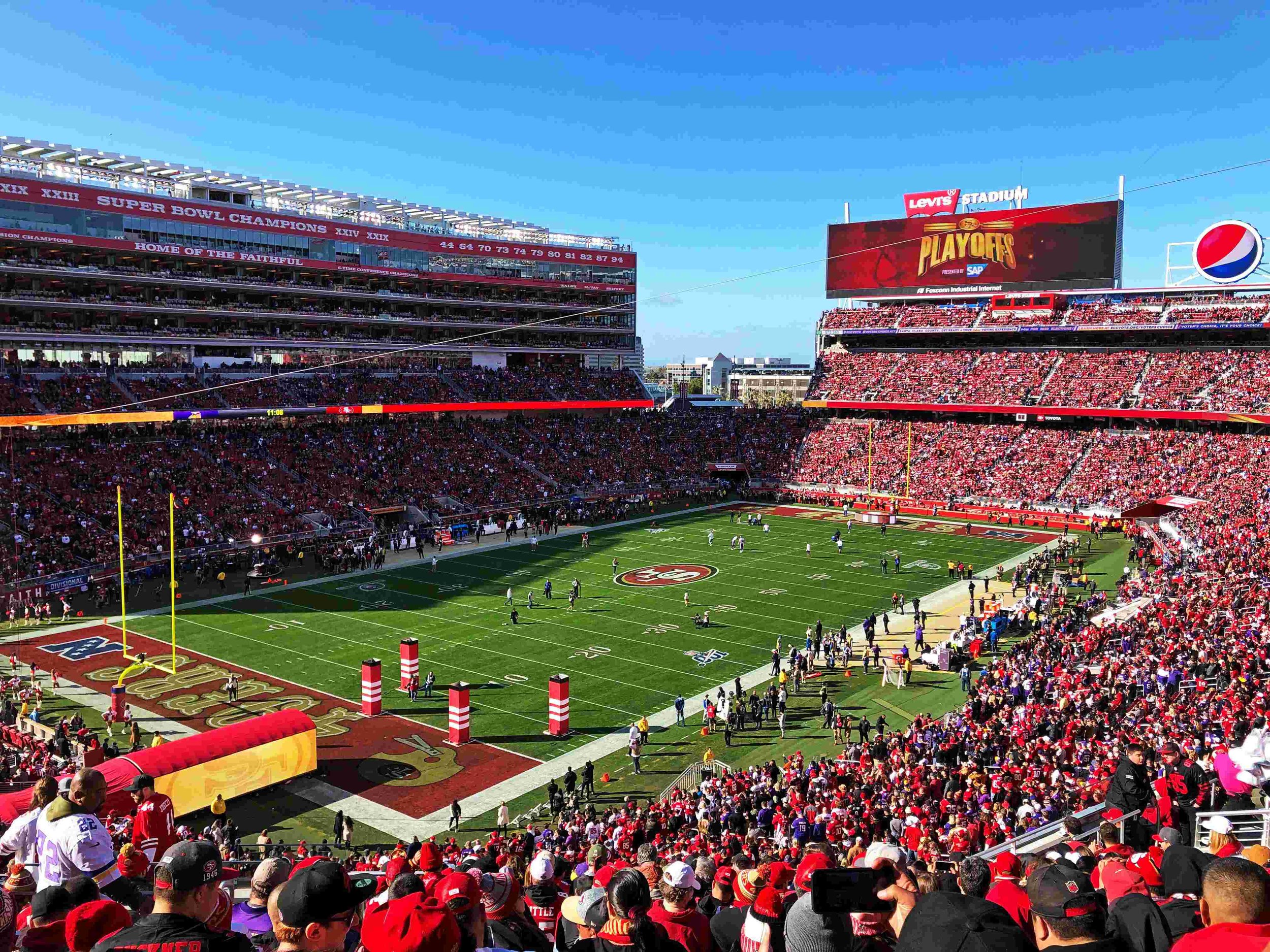 A football stadium filled with fans, with a green field, goalposts, and a large digital scoreboard displaying 'PLAYOFFS' in the background.