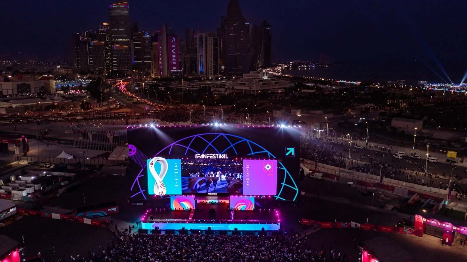 A large outdoor concert stage at night with bright colorful lights, a big screen displaying graphics, and a crowd of people watching, with a city skyline and illuminated buildings in the background.