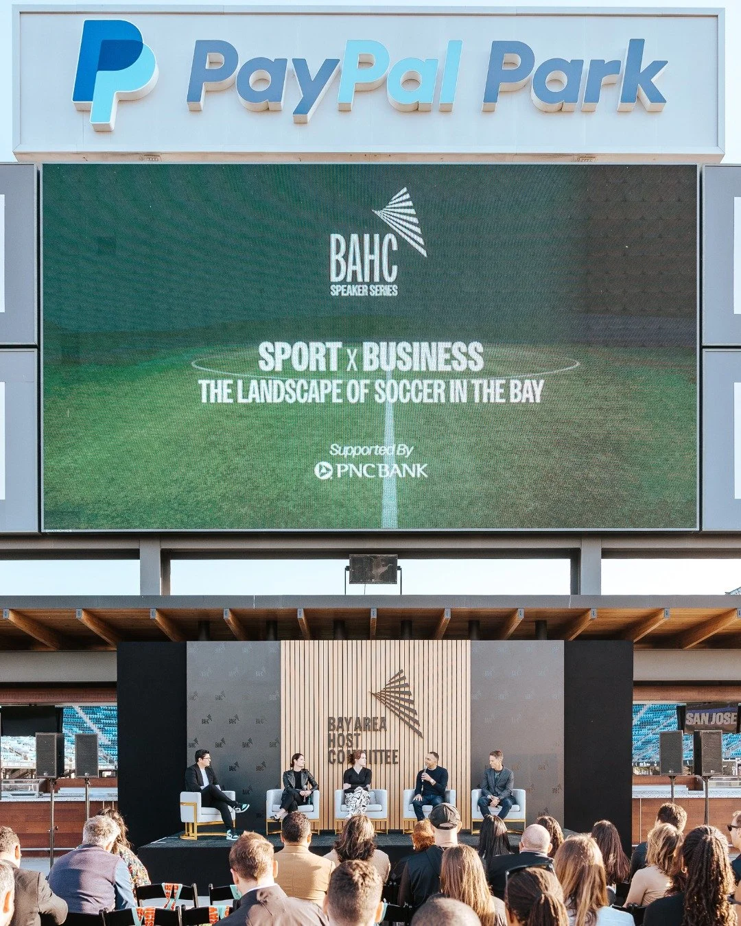An outdoor event at PayPal Park featuring a panel discussion about soccer and business. A large digital screen displays the event name, sponsored by PNC Bank. The stage has four panelists seated in chairs with an audience in front of them.