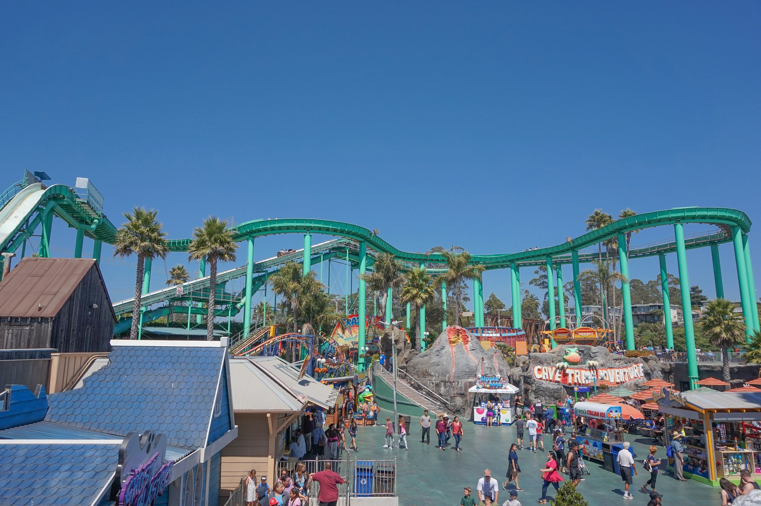A busy amusement park with a roller coaster track painted in teal, surrounded by palm trees, colorful stalls, and many visitors enjoying the sunny day.