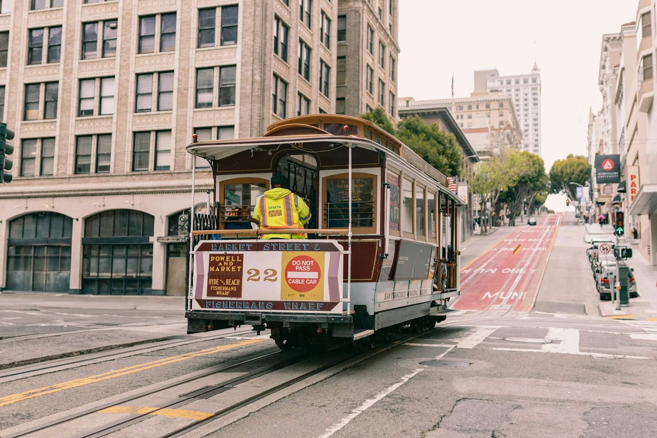 A historic cable car on Market Street in San Francisco, with a driver wearing a reflective vest, city buildings in the background, and overhead wires for the cable car system.