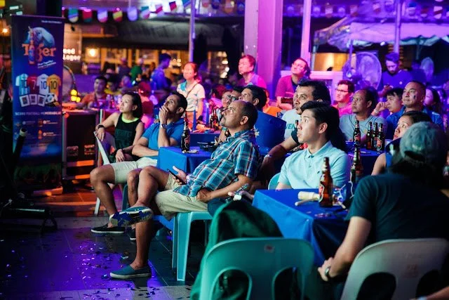 People watching an outdoor entertainment or movie event at night, with colorful lights and drinks on tables.