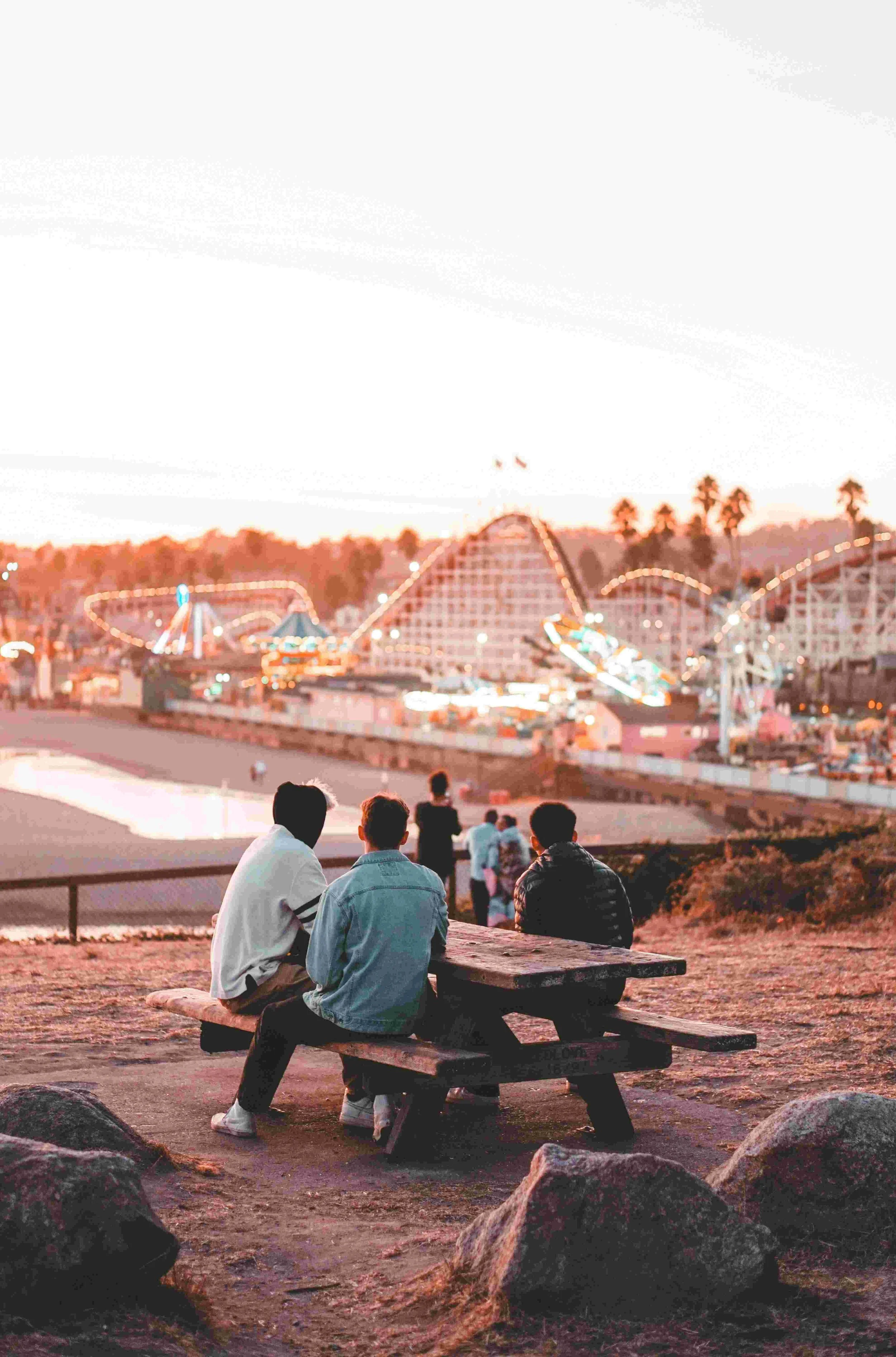 Three young men sitting on a wooden bench at an amusement park during sunset, with a roller coaster and other rides illuminated in the background.