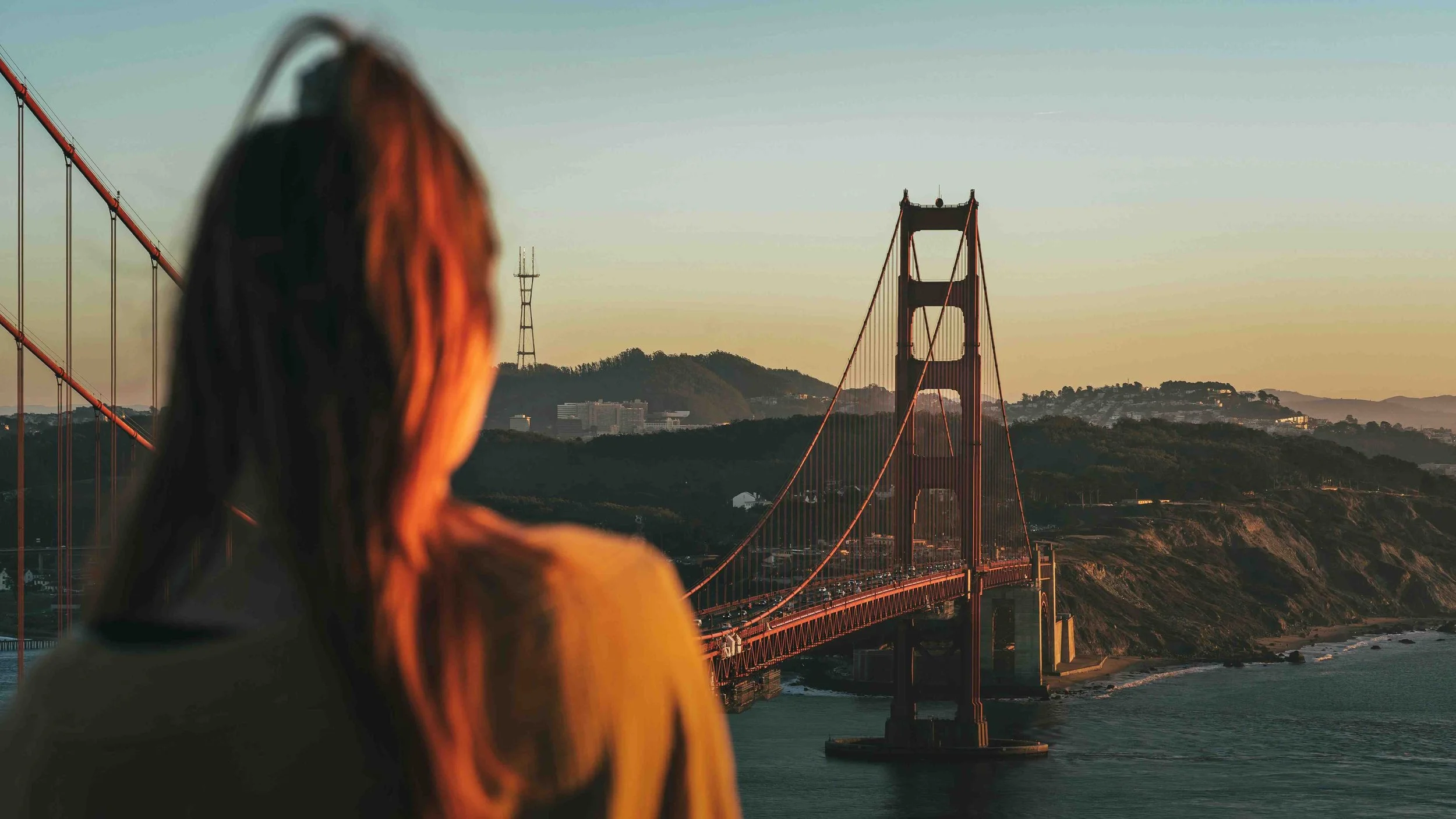A woman with red hair looking at the Golden Gate Bridge during sunset or sunrise in San Francisco.