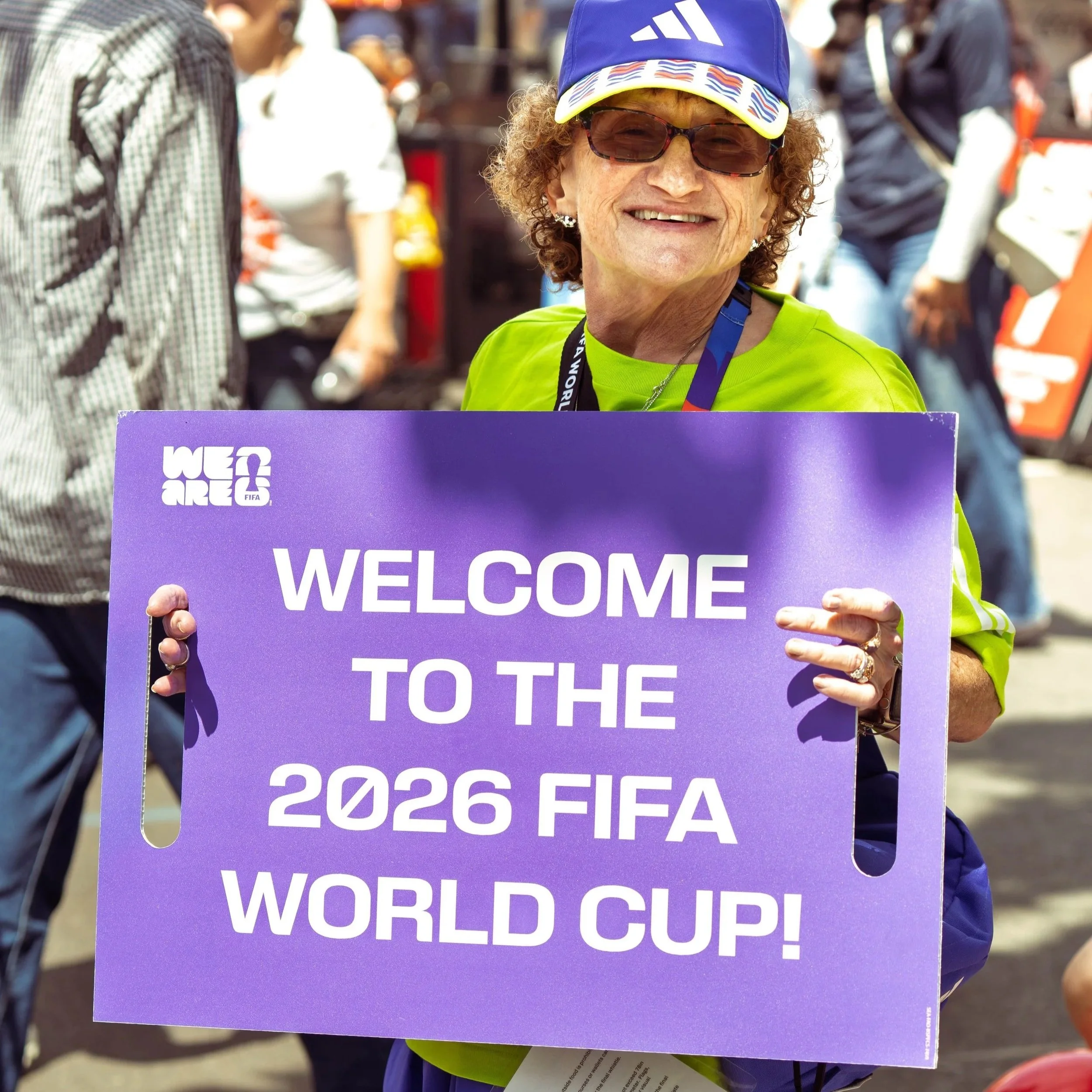 A woman wearing sunglasses and a blue cap holding a purple sign that reads 'WELCOME TO THE 2026 FIFA WORLD CUP' during an outdoor event. The background shows other people and event activities.
