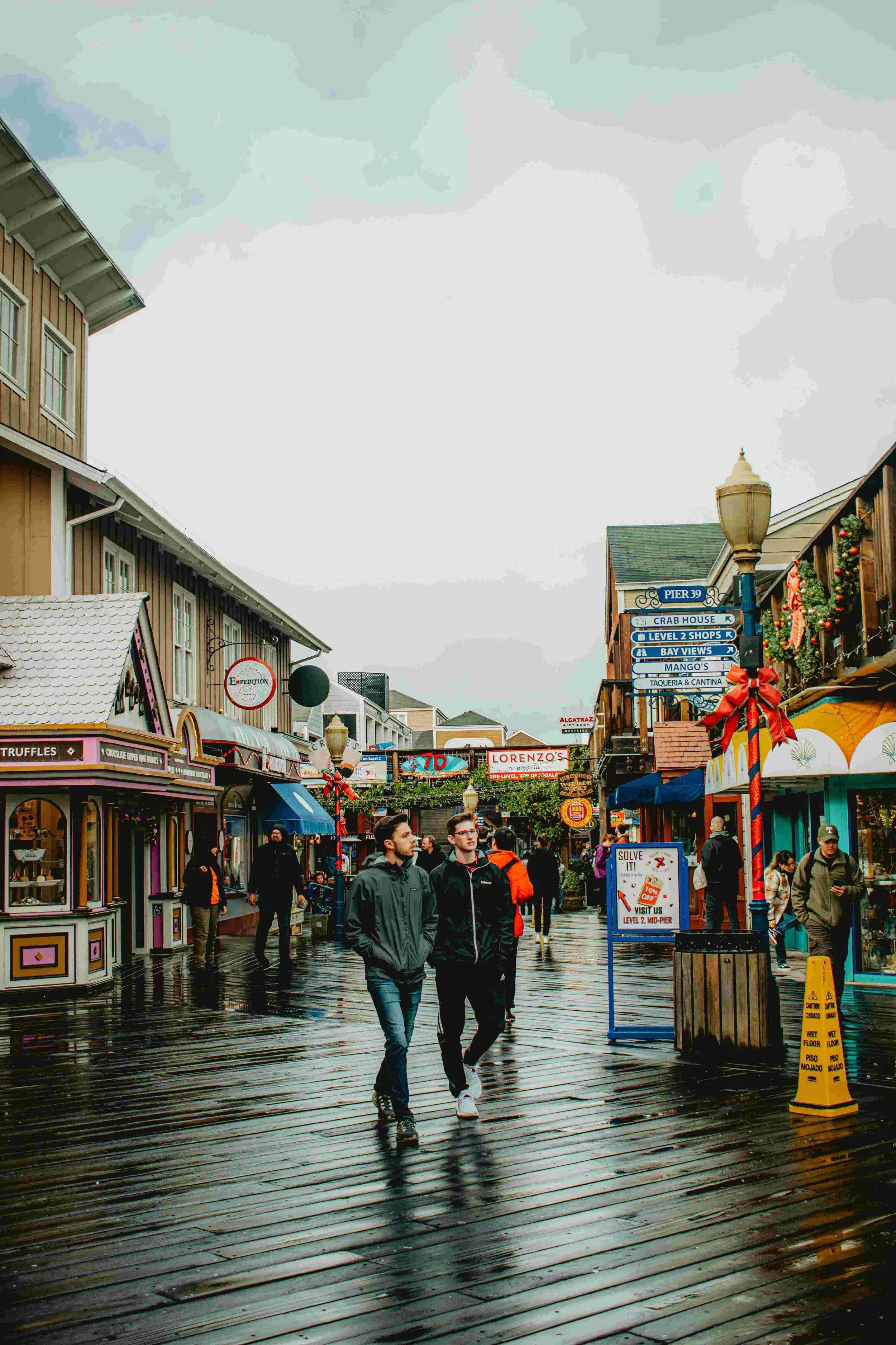 People walking on a wet wooden boardwalk lined with colorful shops and holiday decorations under a cloudy sky.