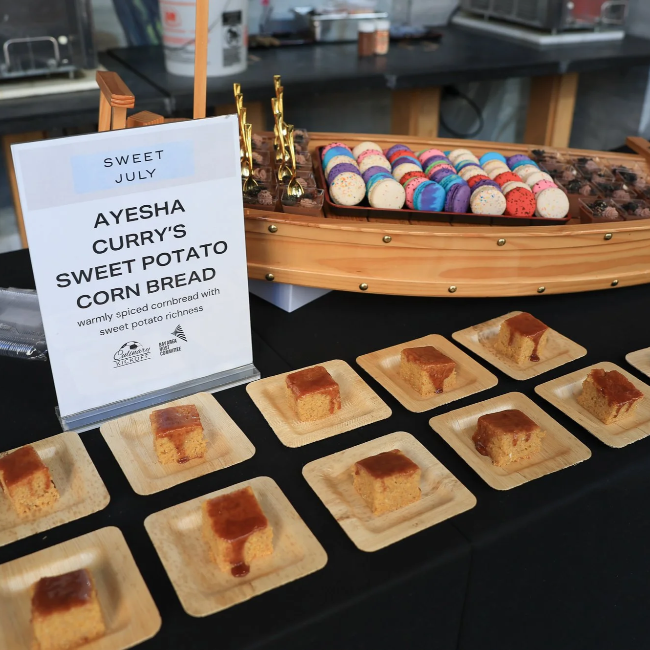 A display of small squares of caramel corn bread topped with caramel sauce arranged on black trays, with a sign describing the sweet potato corn bread at a food event.