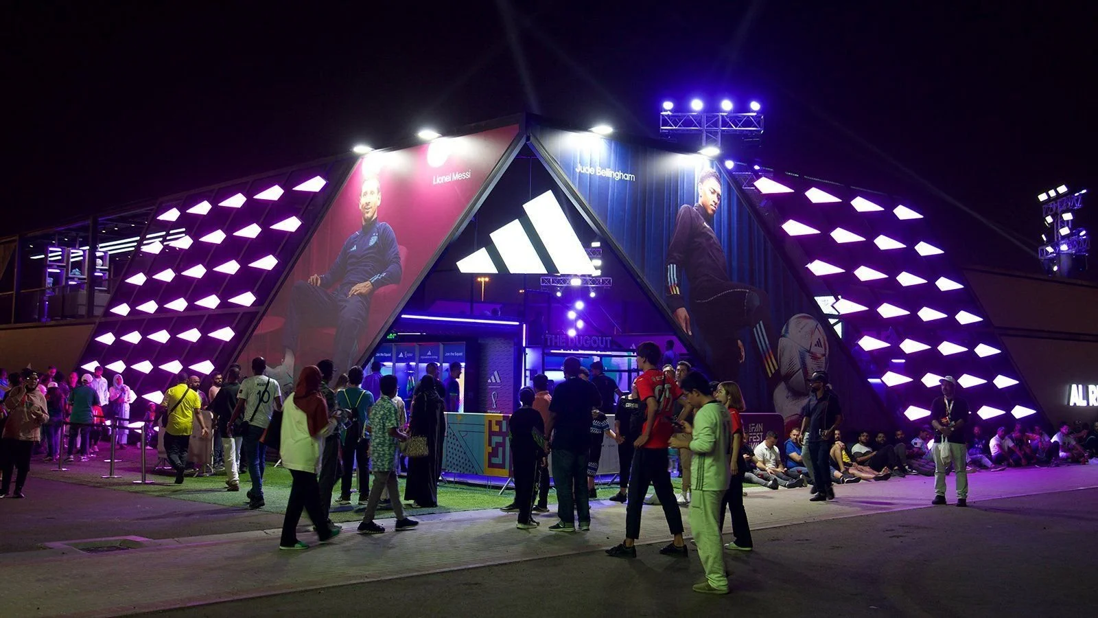 Night scene of an Adidas promotional event with large display screens showing famous football players Lionel Messi and Jude Bellingham, purple and blue lighting, and a crowd of people outside.