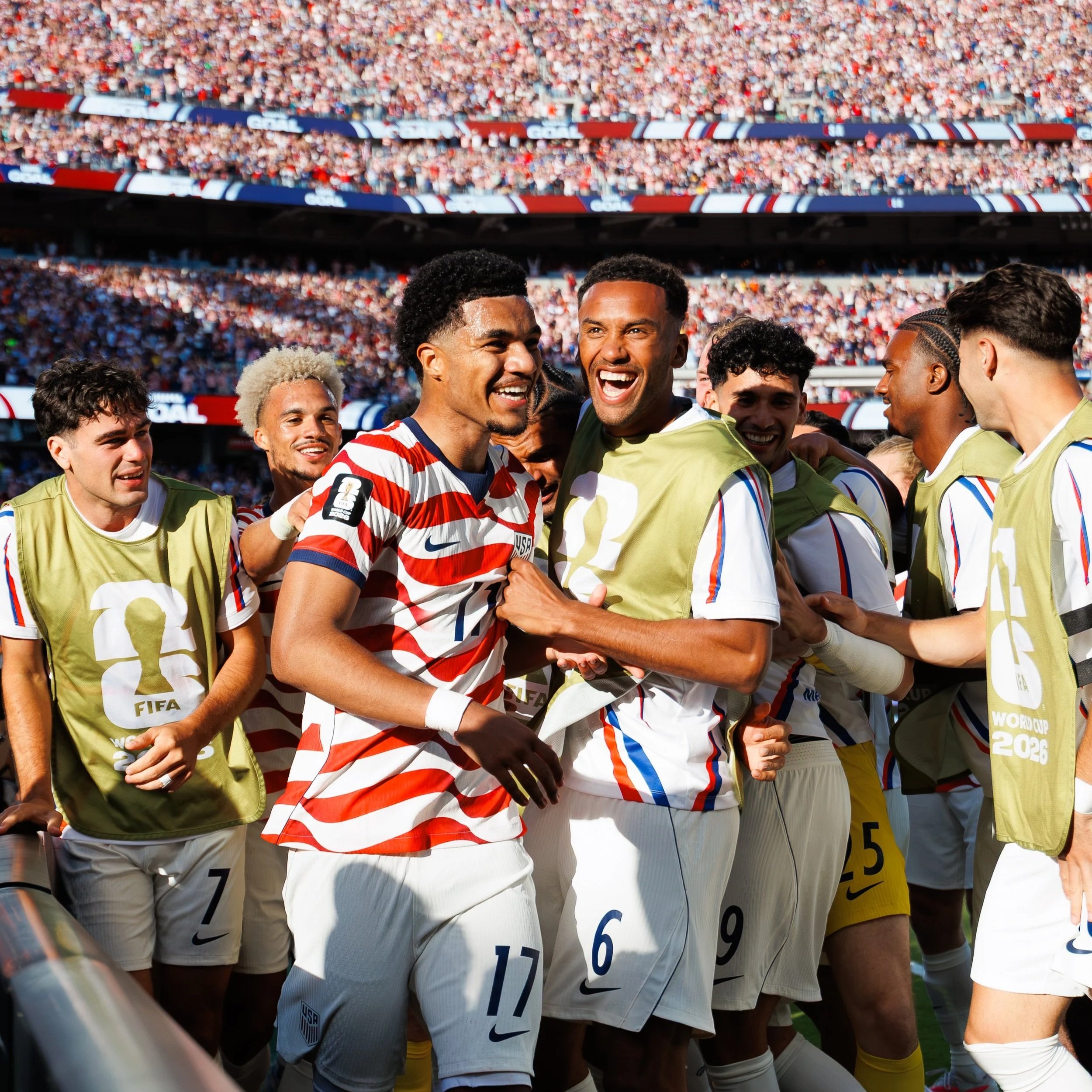 Soccer team members celebrating and smiling on the field during a match, with a large crowd of spectators in the stands.