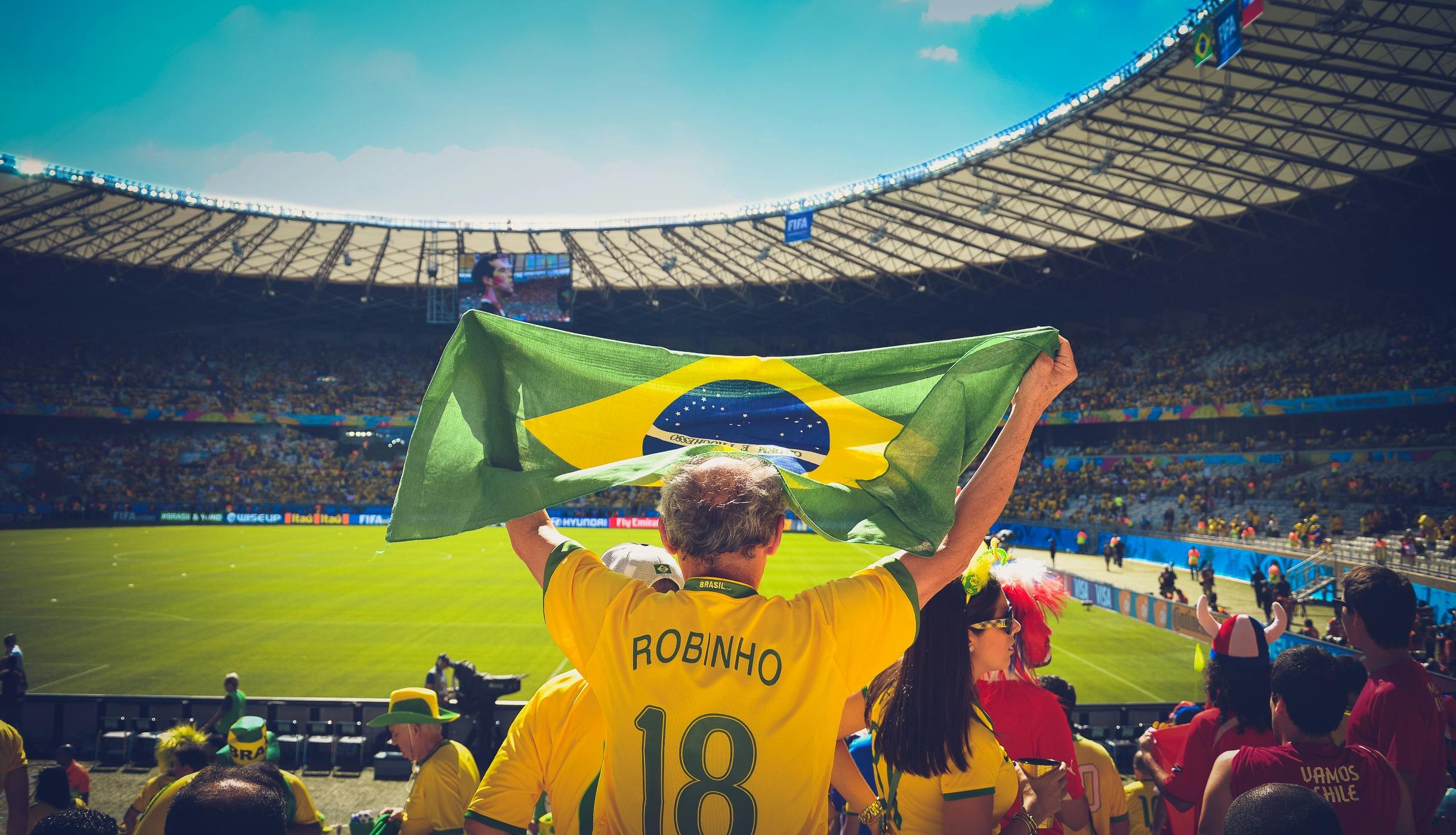 A man wearing a yellow Brazil soccer jersey with 'Robinho' and the number 18, holding a Brazilian flag over his head, in a stadium during a soccer match with fans in festive attire.
