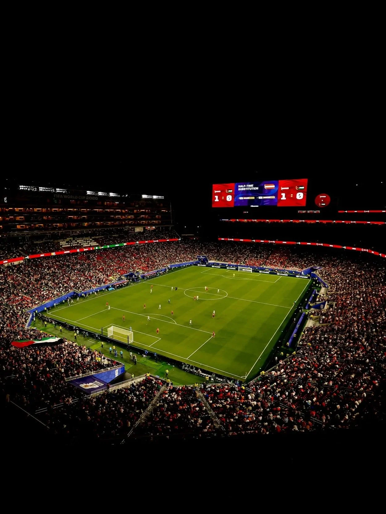 Nighttime photo of a packed soccer stadium during a match, with players on the field, digital scoreboard displaying 1-0, and spectators in the stands.