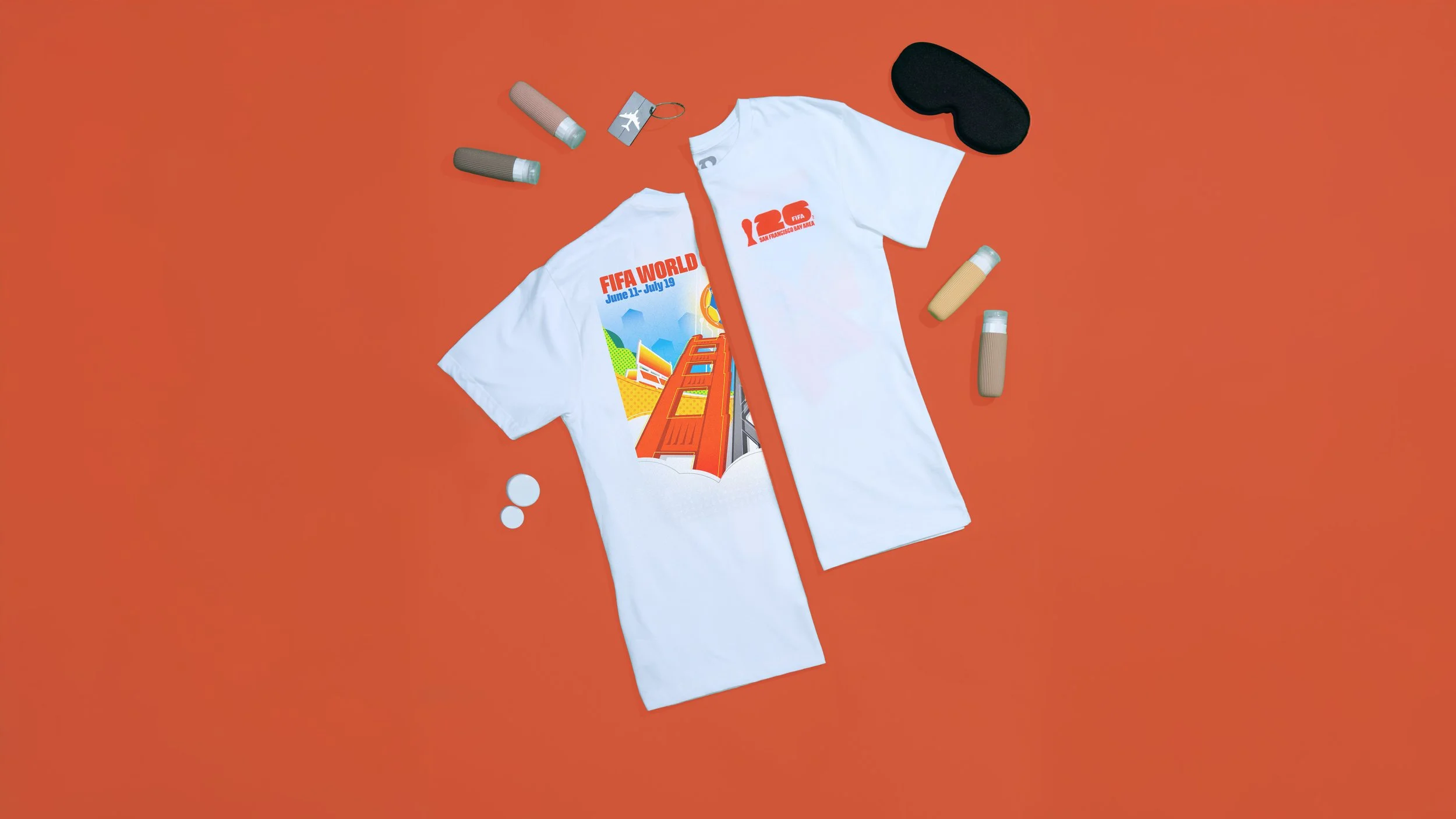 Two white t-shirts stacked on top of each other with a FIFA World Cup graphic on the front, lying on an orange background. Surrounding the shirts are various objects including a sleep mask, small bottles, a keychain, and a couple of white pills.