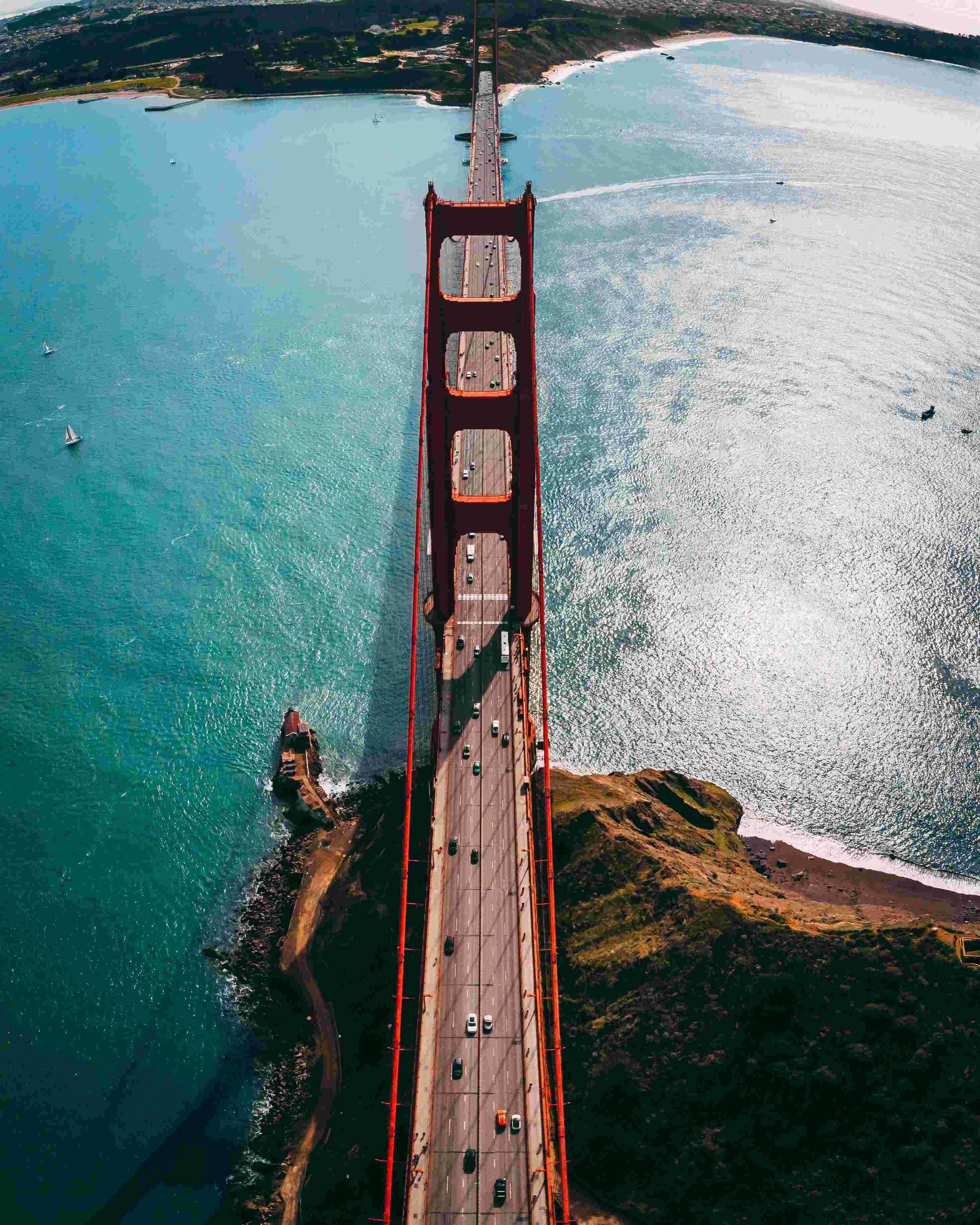 Aerial view of the Golden Gate Bridge with cars crossing, spanning over water with sailboats and a coastline visible.