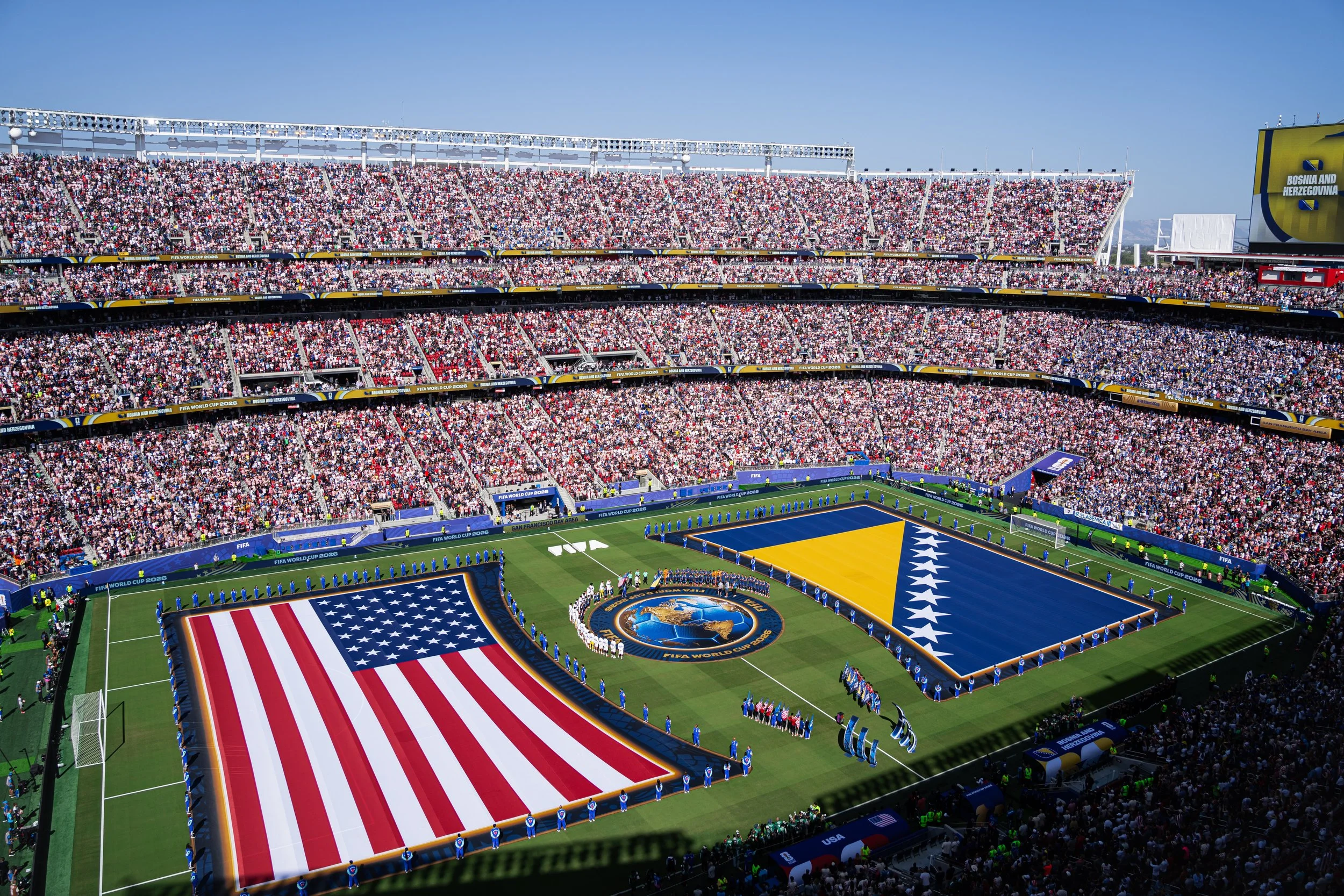 A packed soccer stadium during a FIFA World Cup match, with the national flag of the United States displayed on the field and the crowd in the stands.