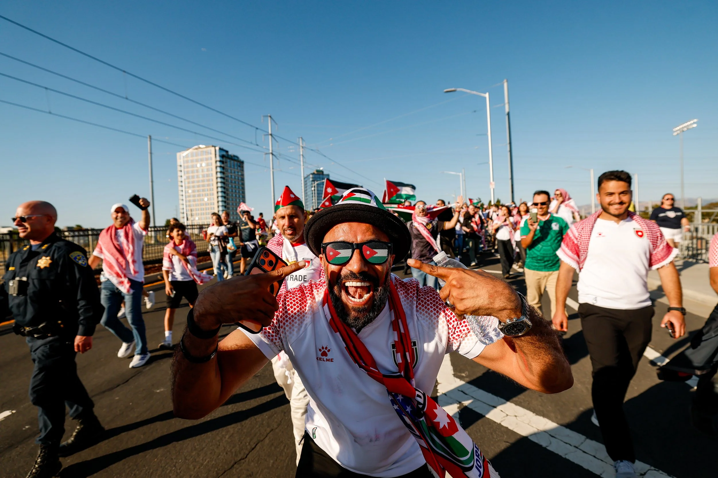 A group of people outdoors celebrating, with some wearing flags and others holding Brazilian flags, along with a police officer and city buildings in the background.