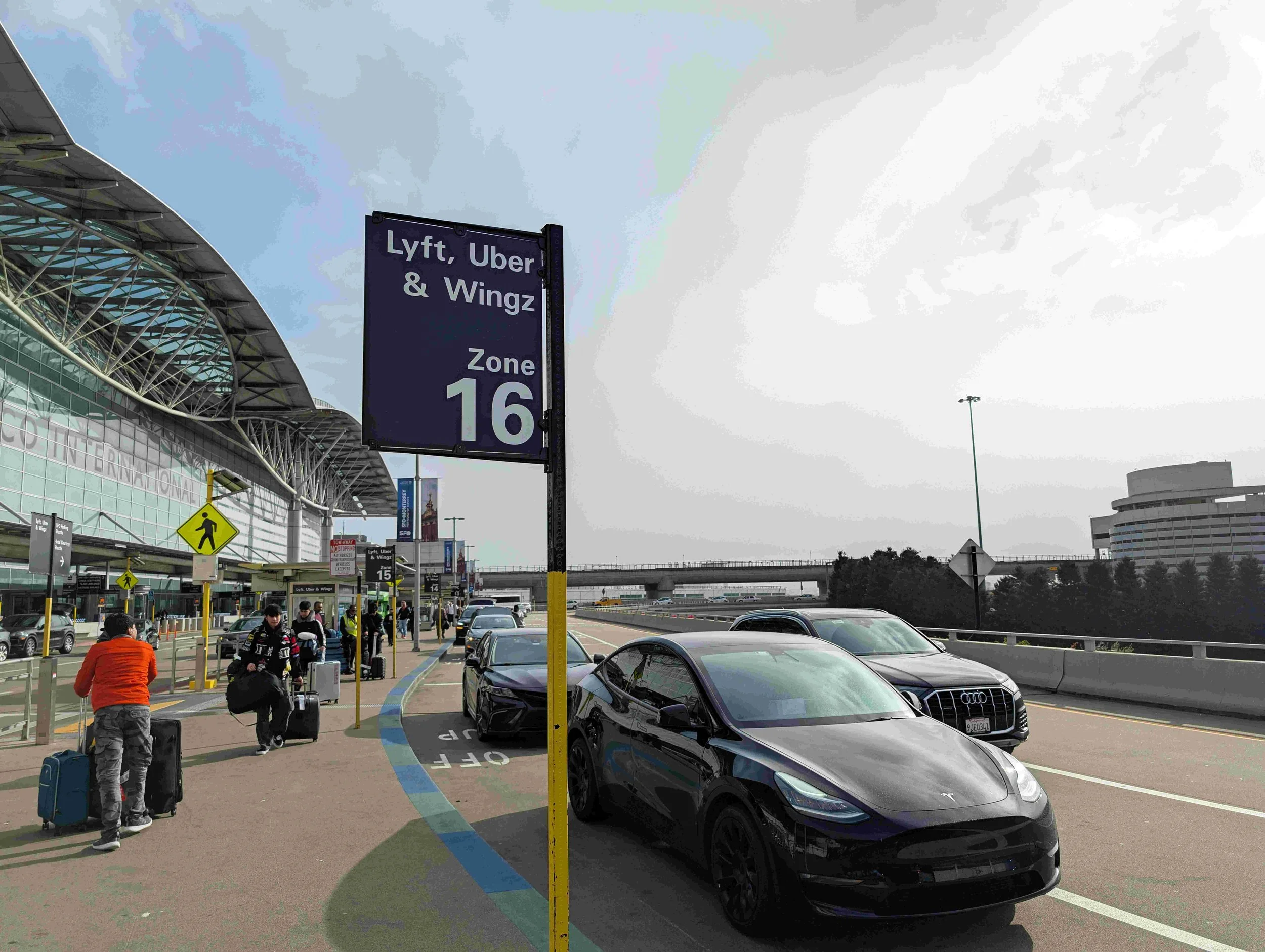 Passengers with luggage walking outside an airport terminal near parked cars, with a sign indicating Zone 16 for Lyft, Uber, and Wingz pickups.