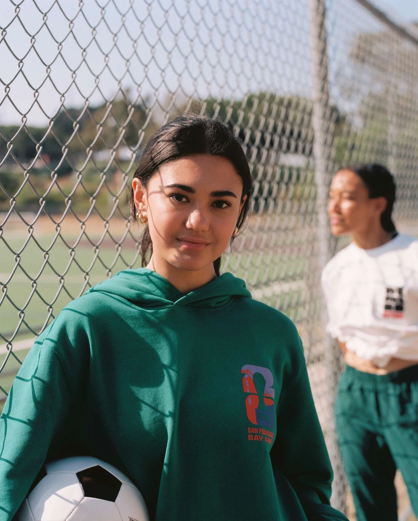 Young woman in green hoodie holding soccer ball, standing by a chain-link fence on a soccer field, with a woman in white shirt blurred in the background.