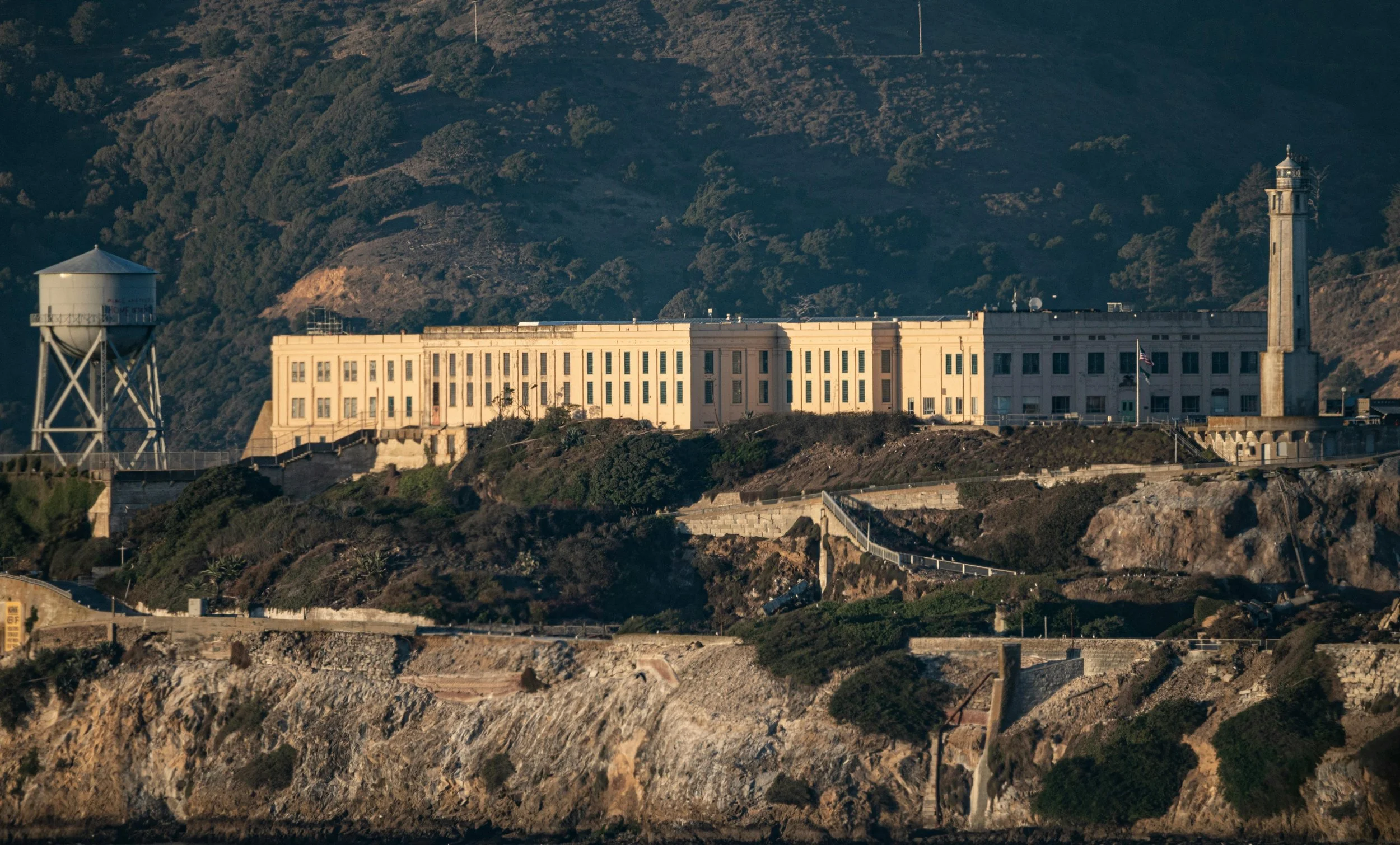 The historic Alcatraz Island prison building with a lighthouse on the right and a water tower on the left, set against forested hills.