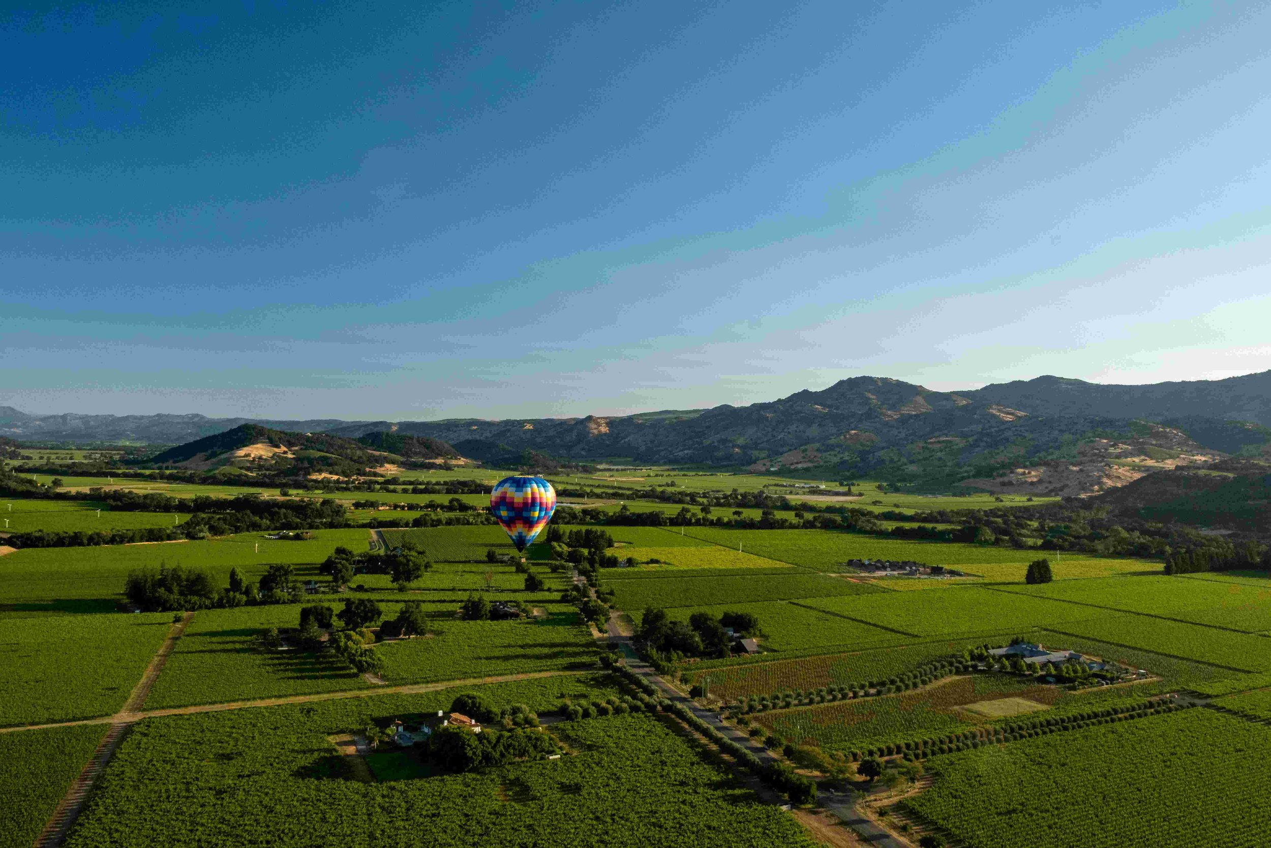 A colorful hot air balloon floating over a lush green rural landscape with fields, trees, and mountains in the background under a blue sky.