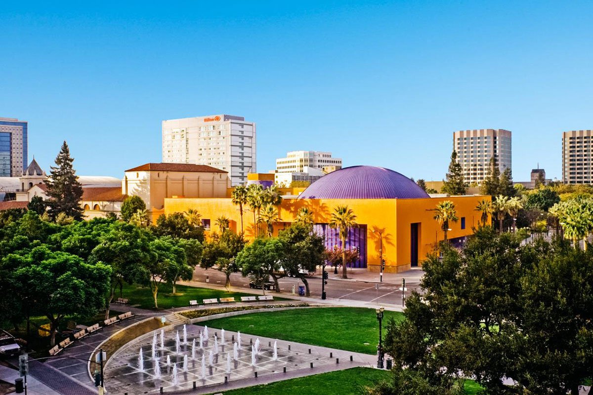 Colorful city park with a yellow building with a purple dome, surrounded by green trees, benches, and a fountain, with tall buildings in the background under a clear blue sky.