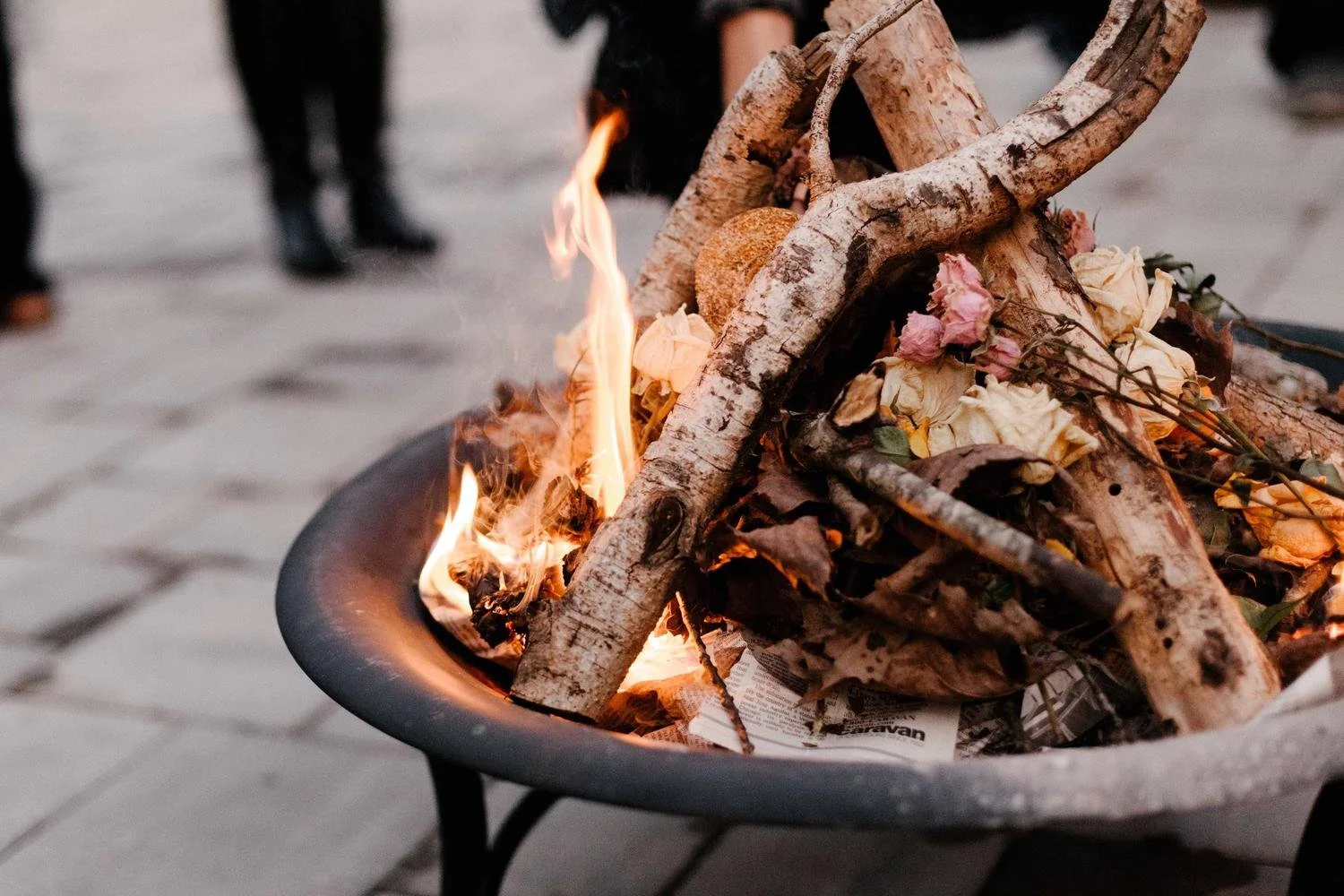 A fire burning in a black fire pit with some wooden logs and flower petals on top, set against a background of blurry people and pavement.