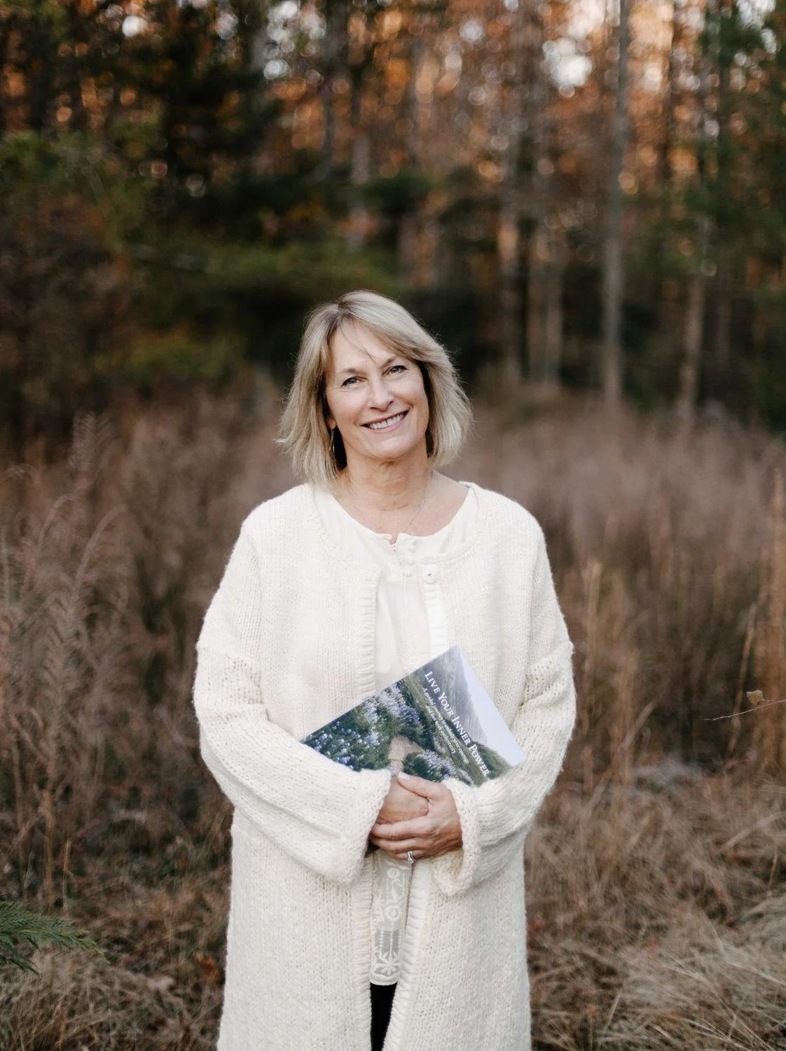 A smiling middle-aged woman with blonde hair stands outdoors in a wooded area during autumn, holding a magazine titled "Live Your Inner Power." She wears a cream-colored cardigan over a white blouse.