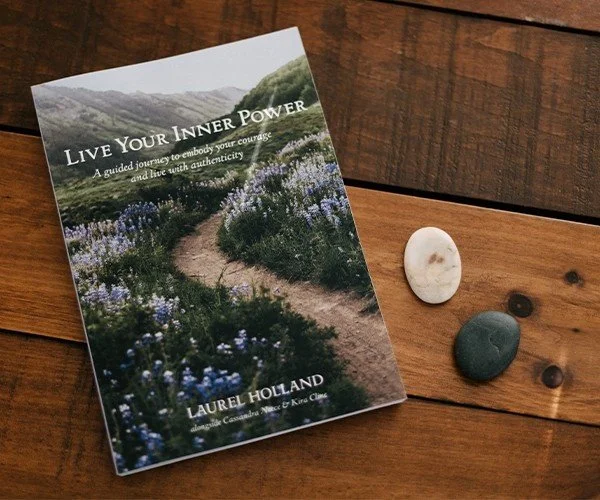 A book titled "Live Your Inner Power" by Laurel Holland, placed on a wooden surface next to three stones and three small dark pebbles. The book features a scenic cover image of a winding path through a field of purple flowers and greenery.